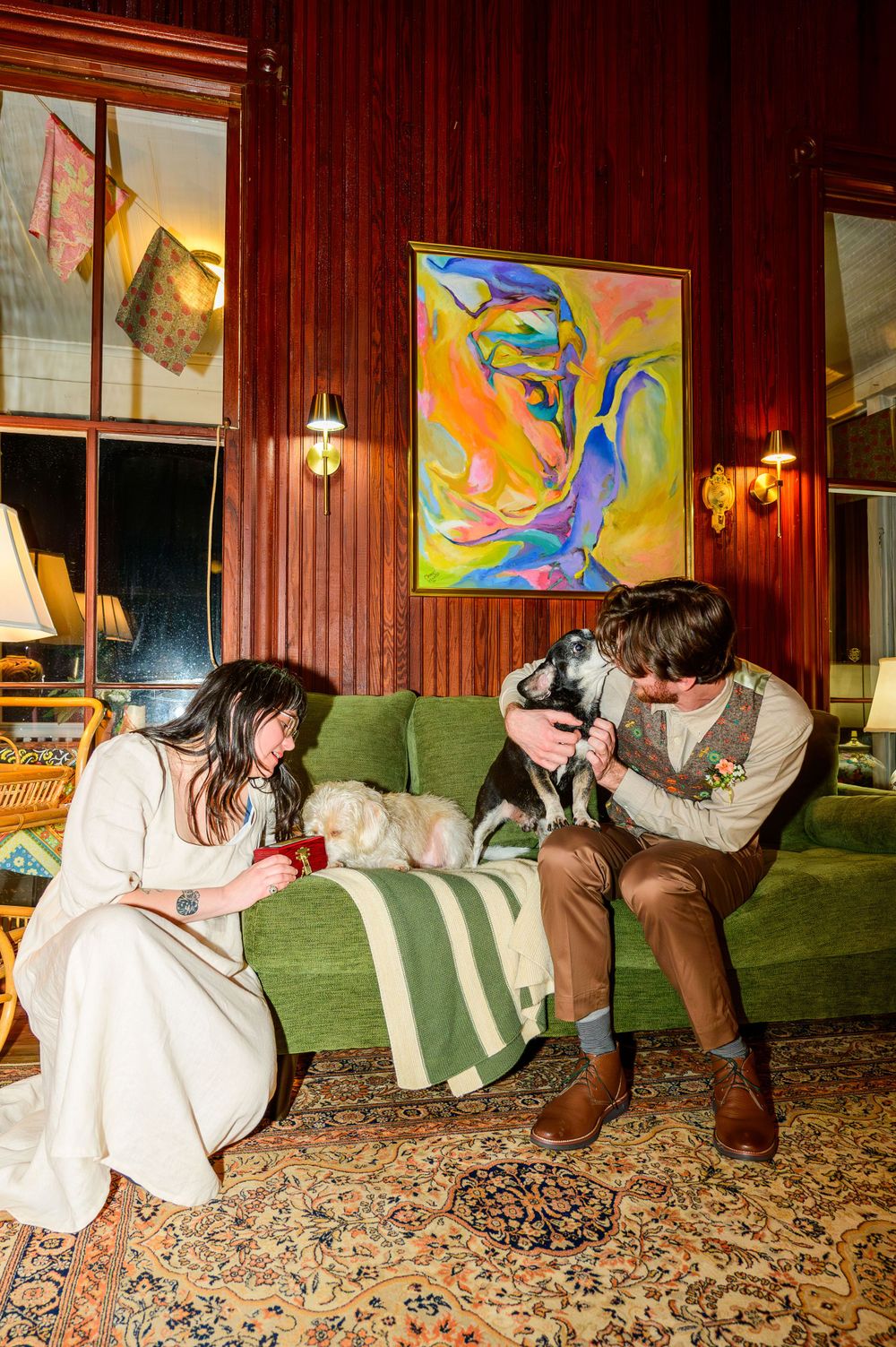 A couple sits on a green couch in a rustic living room with wood paneling and colorful abstract art on the wall.