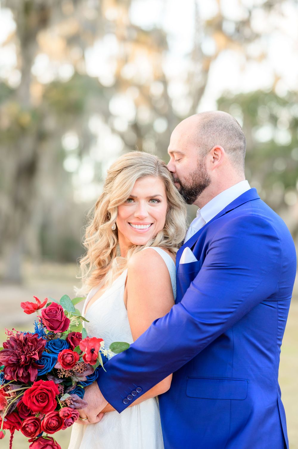 Wedding couple embracing outdoors with vibrant red bouquet against backdrop of Spanish moss trees.