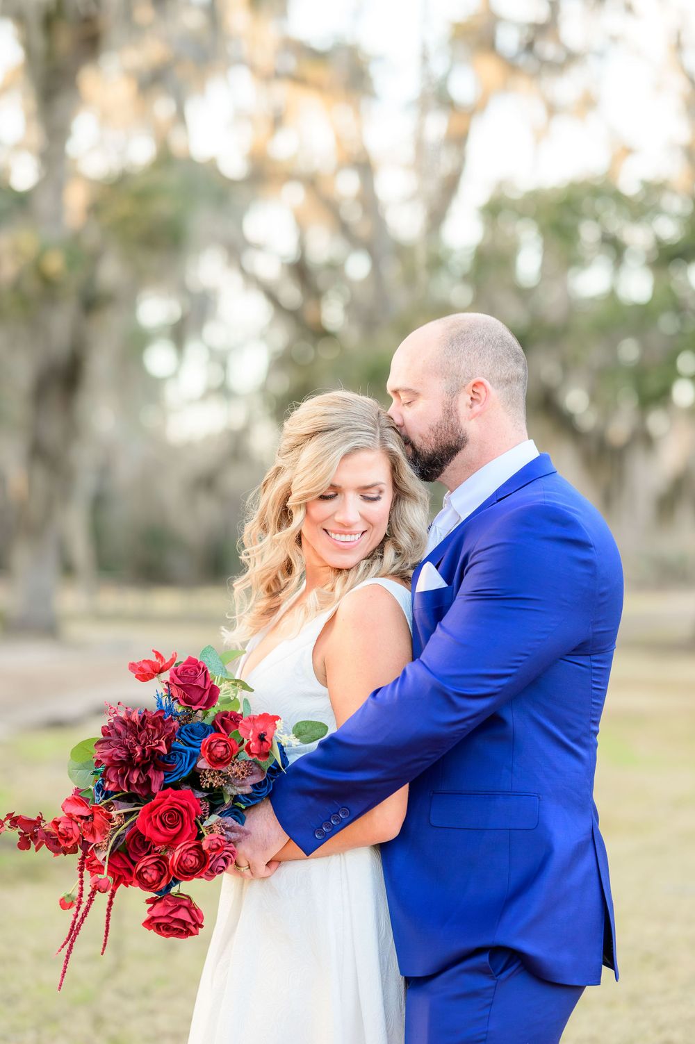 Romantic wedding portrait with bride holding red rose bouquet while groom embraces her.