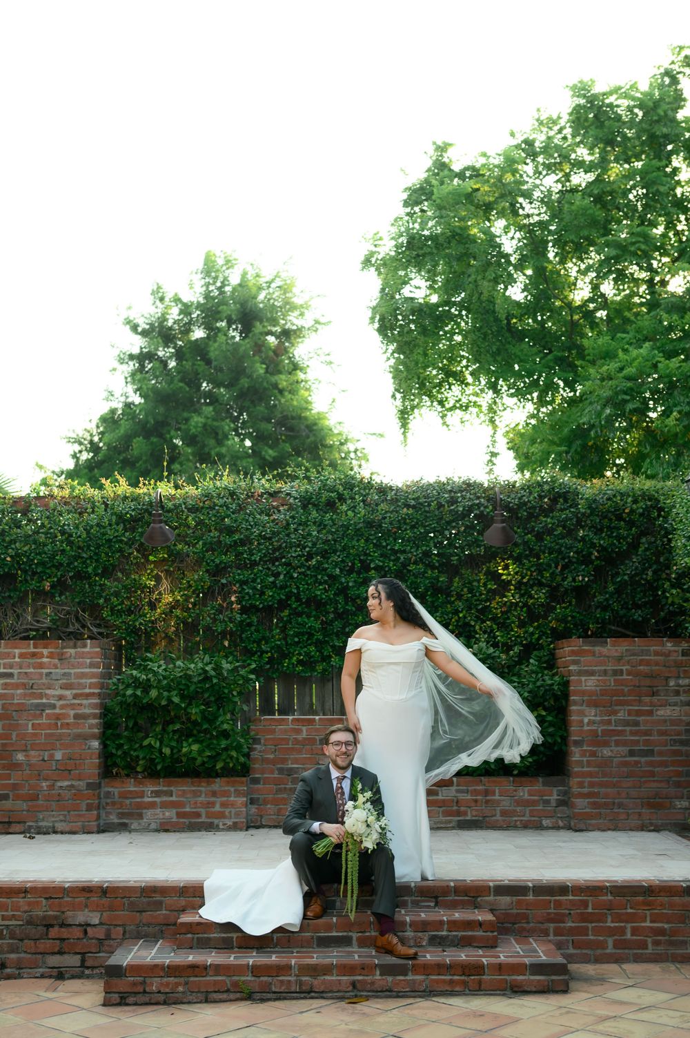 Bride in white gown and veil stands on brick steps beside seated groom holding bouquet, lush green hedge backdrop.