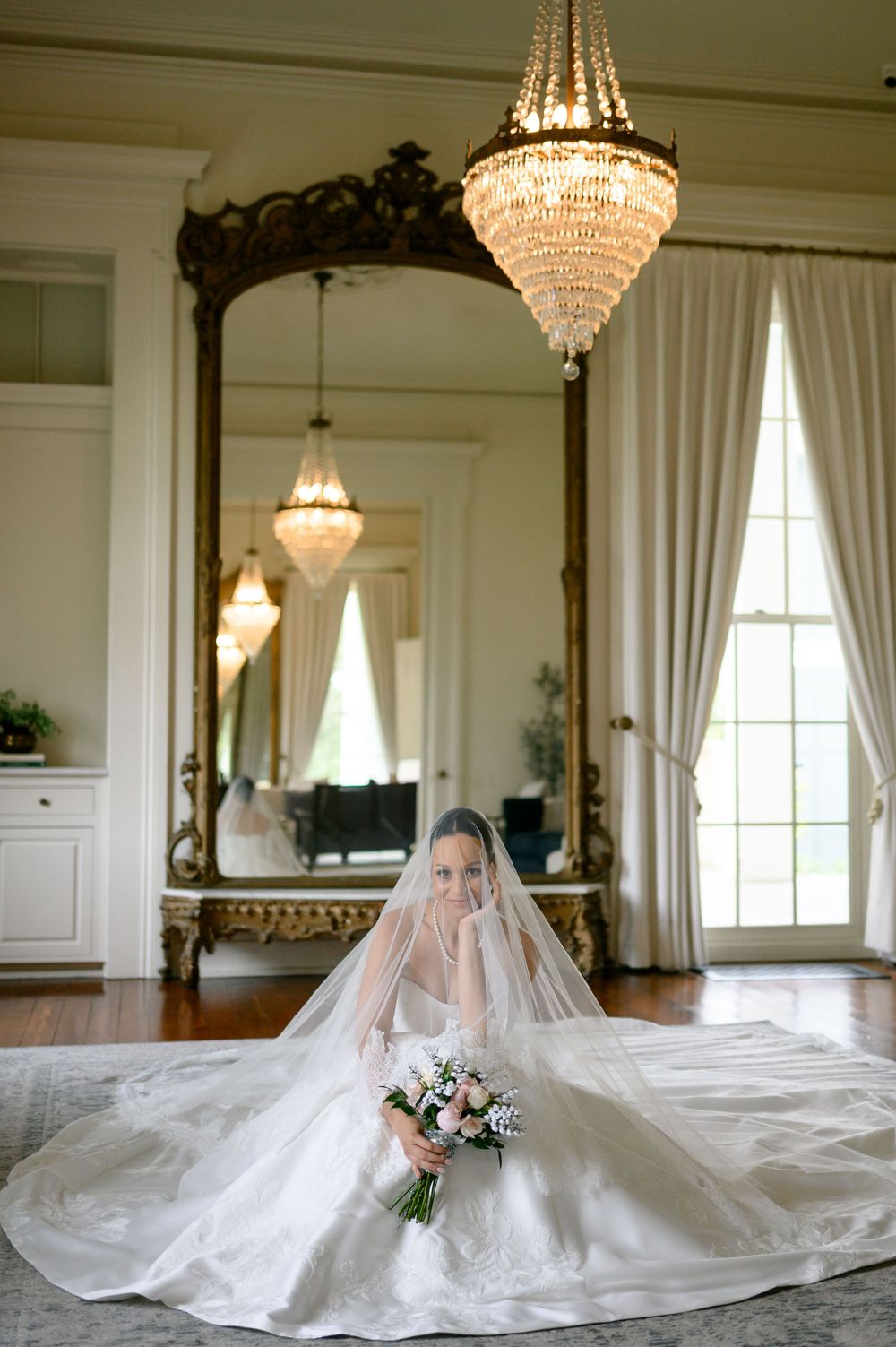 Bride in white lace gown and veil sits on floor holding bouquet under crystal chandelier with ornate gold mirror.