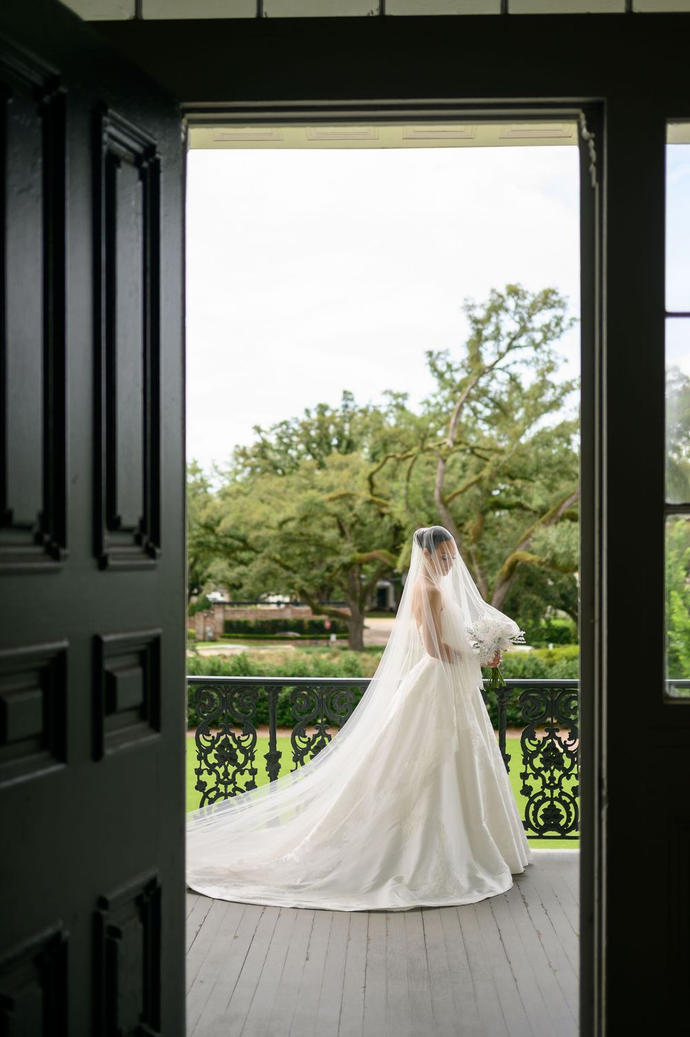 Bride in a cathedral-train wedding gown and veil stands on a balcony framed by dark ornate doors.