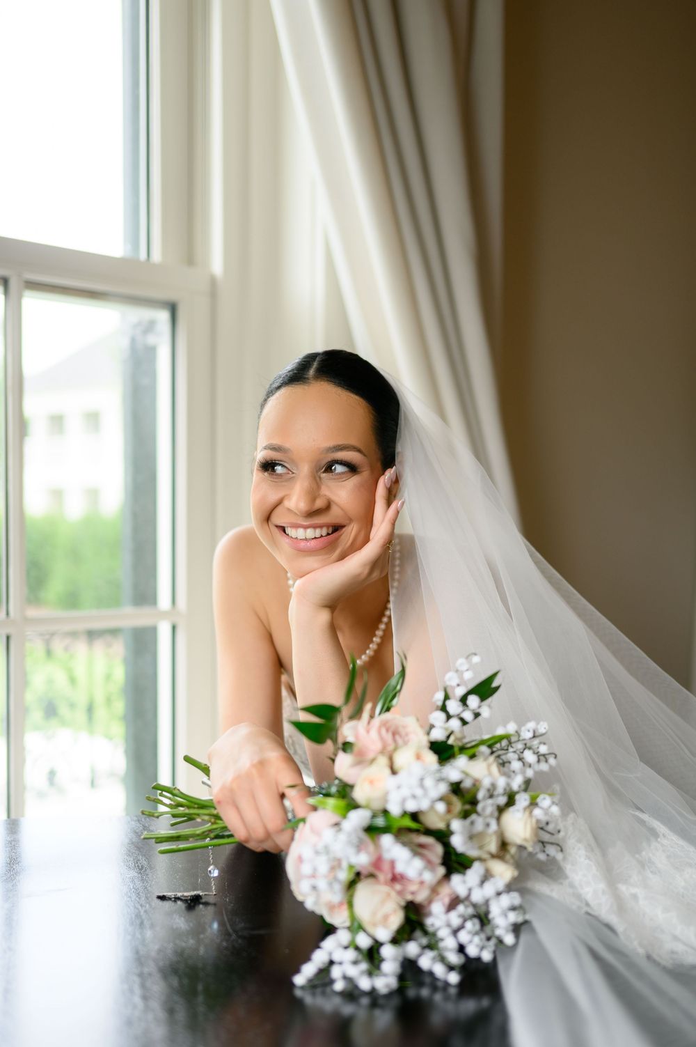 Smiling bride in white veil and pearl necklace leaning on table near window with bridal bouquet of pink and white flowers.