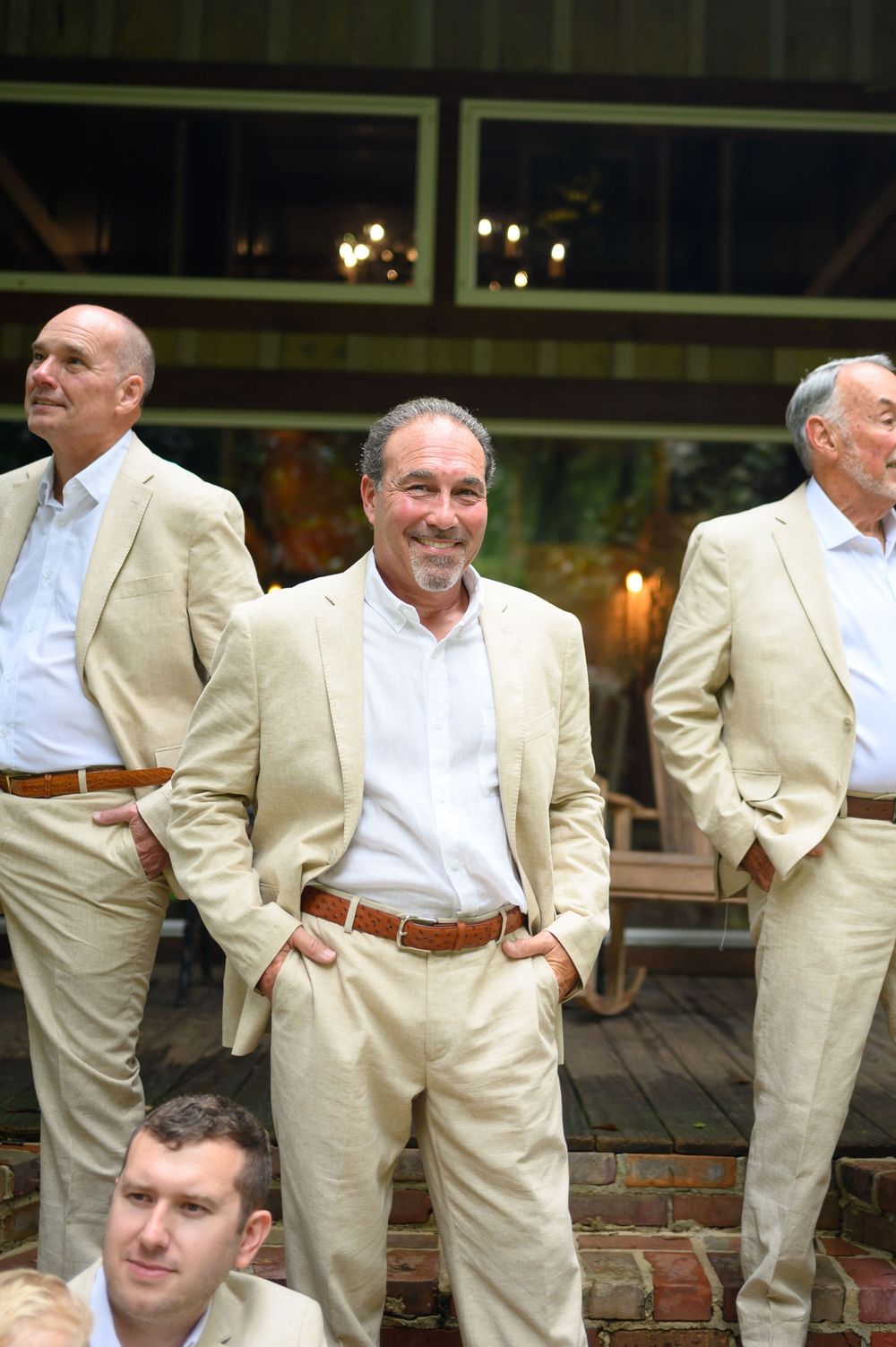 Men in matching tan suits with brown belts stand outdoors at a formal event, smiling and relaxed.