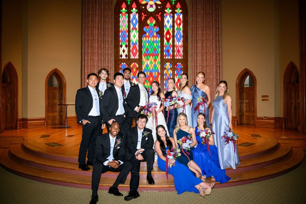 Prom group posing in elegant church with colorful stained glass windows, wearing formal gowns and tuxedos.