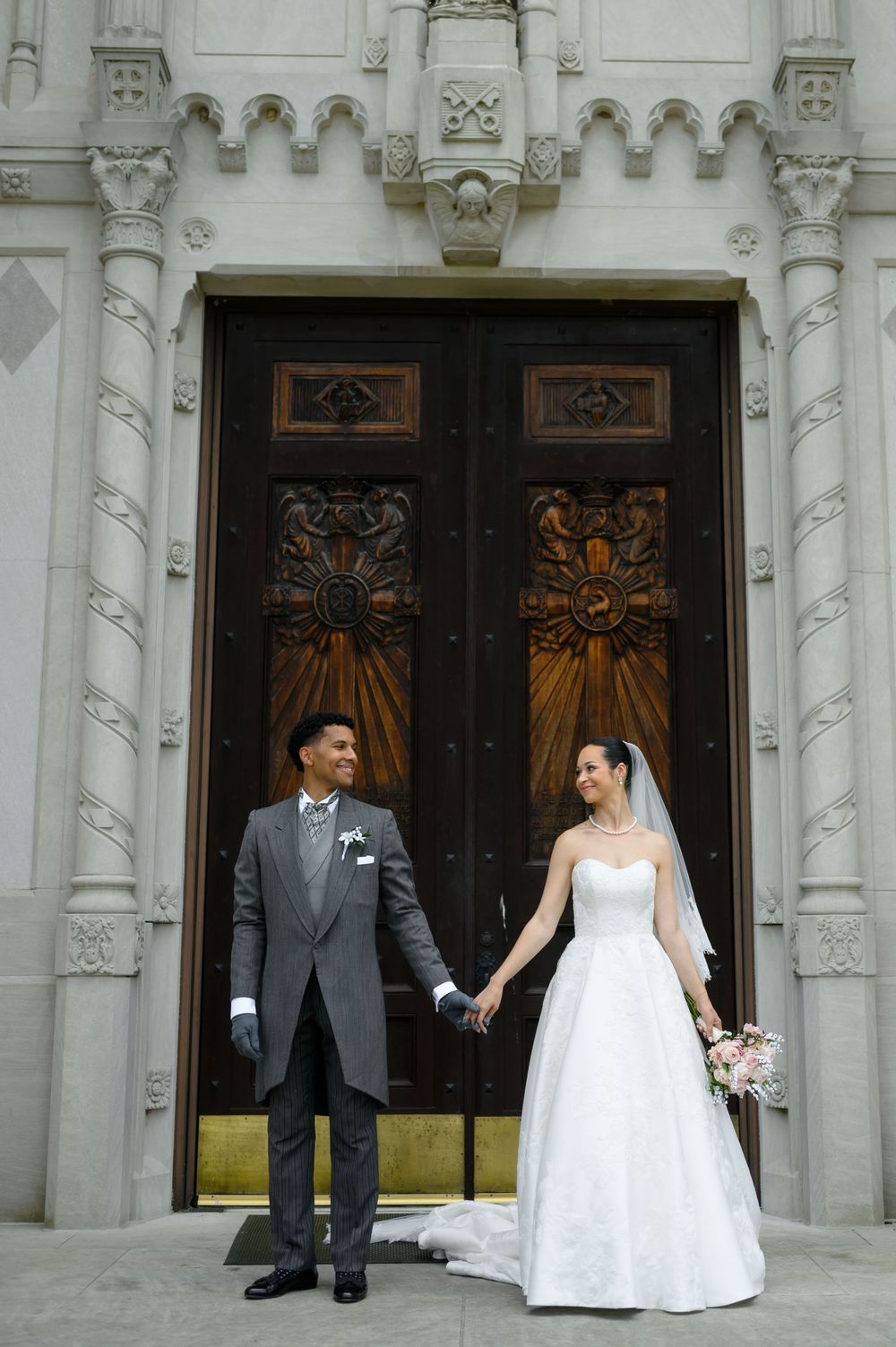 Bride and groom hold hands before ornate church doors, she in white gown with veil, he in gray suit.
