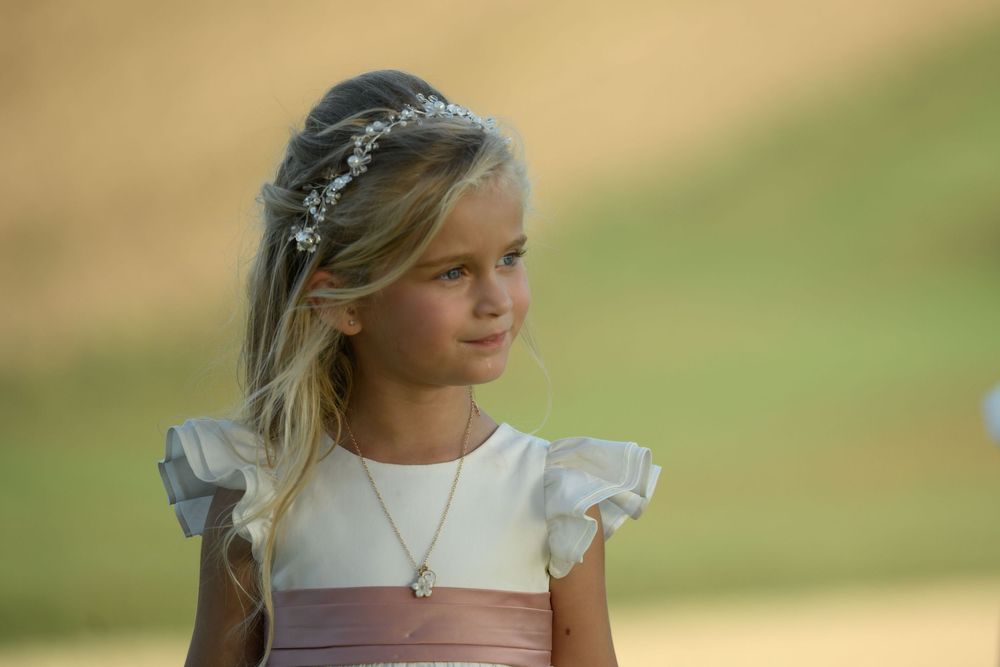 Young flower girl in white dress with pink sash and floral crown, wearing a delicate necklace at an outdoor event.