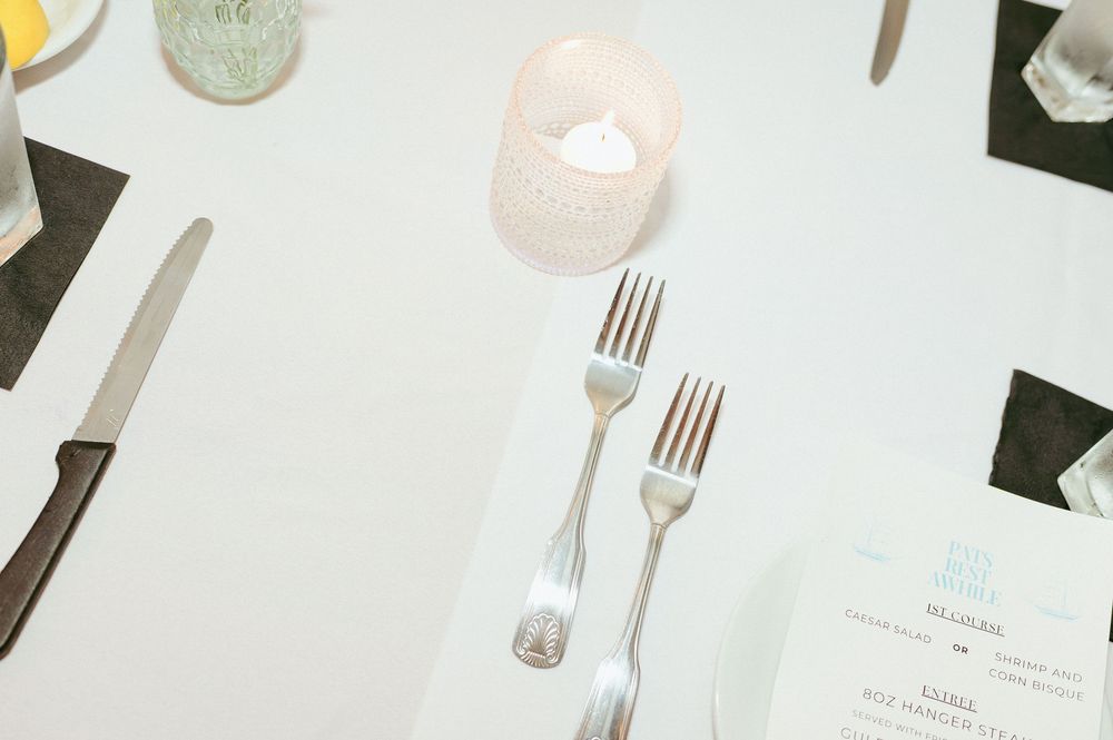 Minimalist table setting with silver forks and glowing votive candle on white tablecloth.
