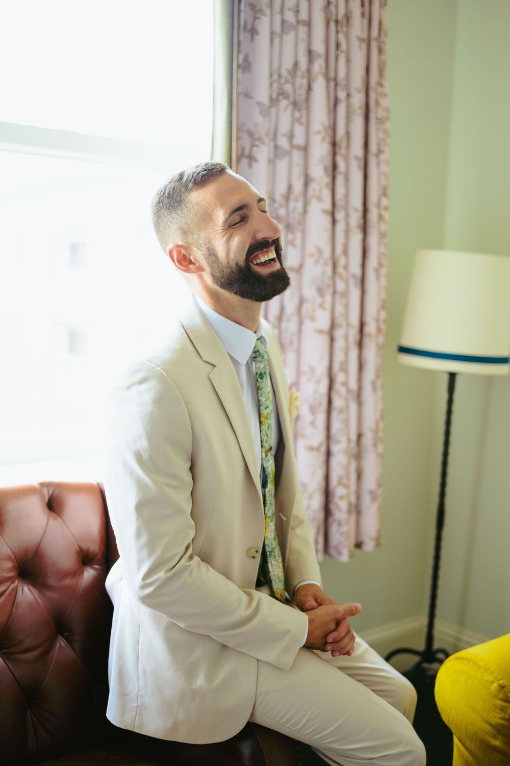 Four sequential photos of a groom in a white suit laughing while seated on a couch in a room with green walls.