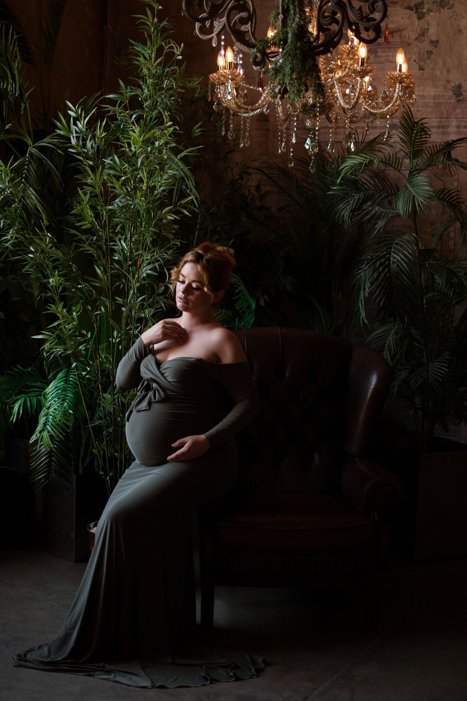 Dramatic maternity portrait in dark room with chandelier and lush greenery.