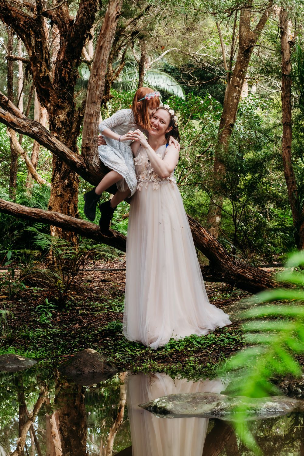 Wedding couple posing playfully by reflective pond in enchanted forest setting.