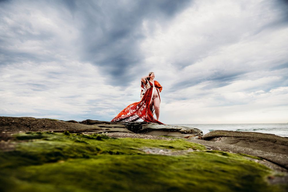 Dramatic coastal wedding photos with red dress against cloudy sky and rocky cliffs.