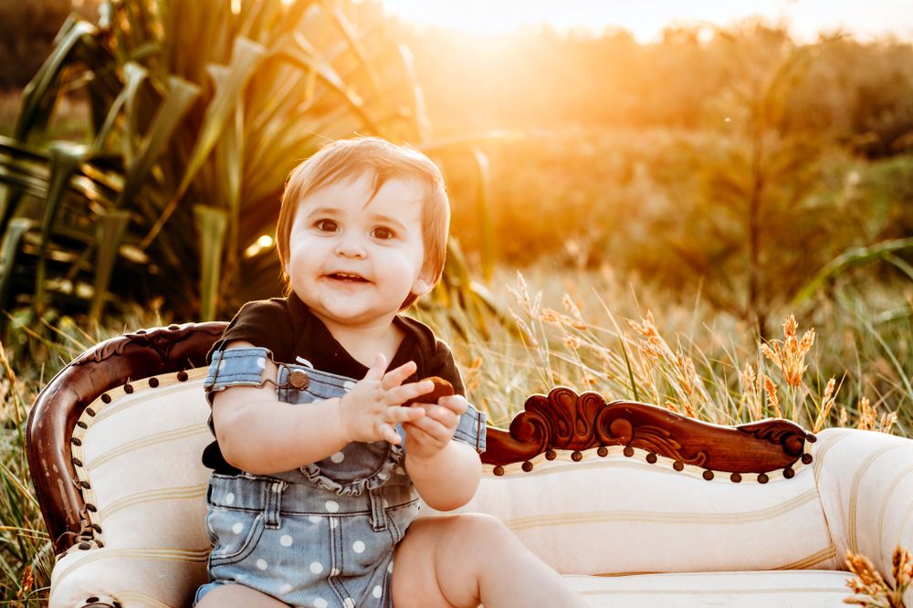 Baby in denim overalls sits on vintage chair during golden hour sunset photoshoot.