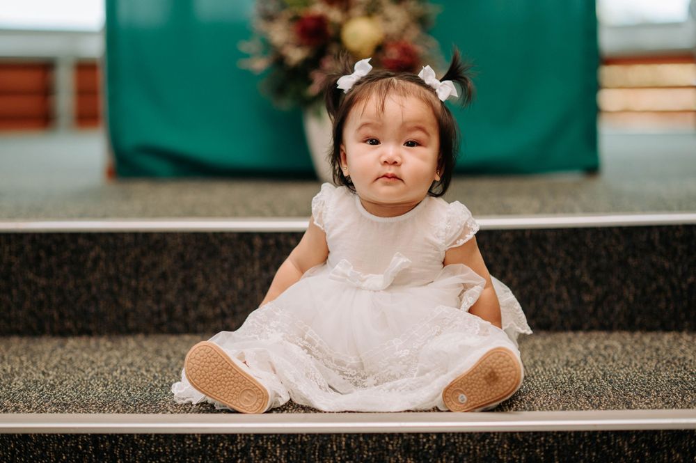 Baby in white lace dress sitting on grey steps with a hair accessory.
