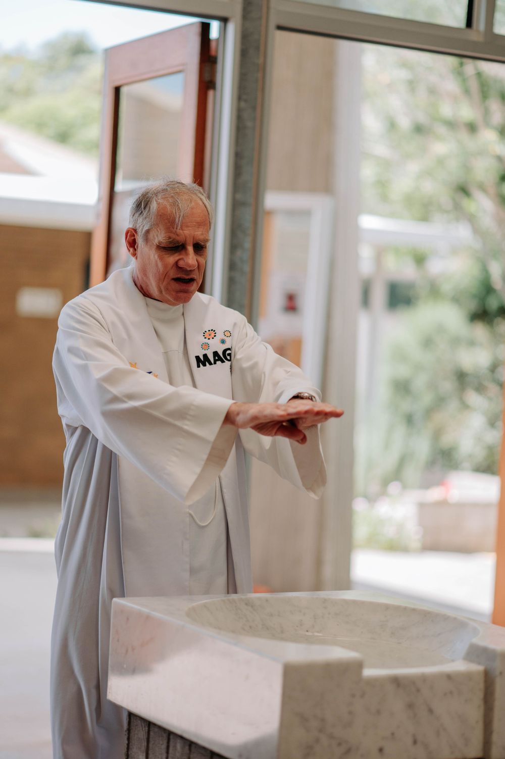 Clergy member in white robe performing ritual at marble baptismal font.