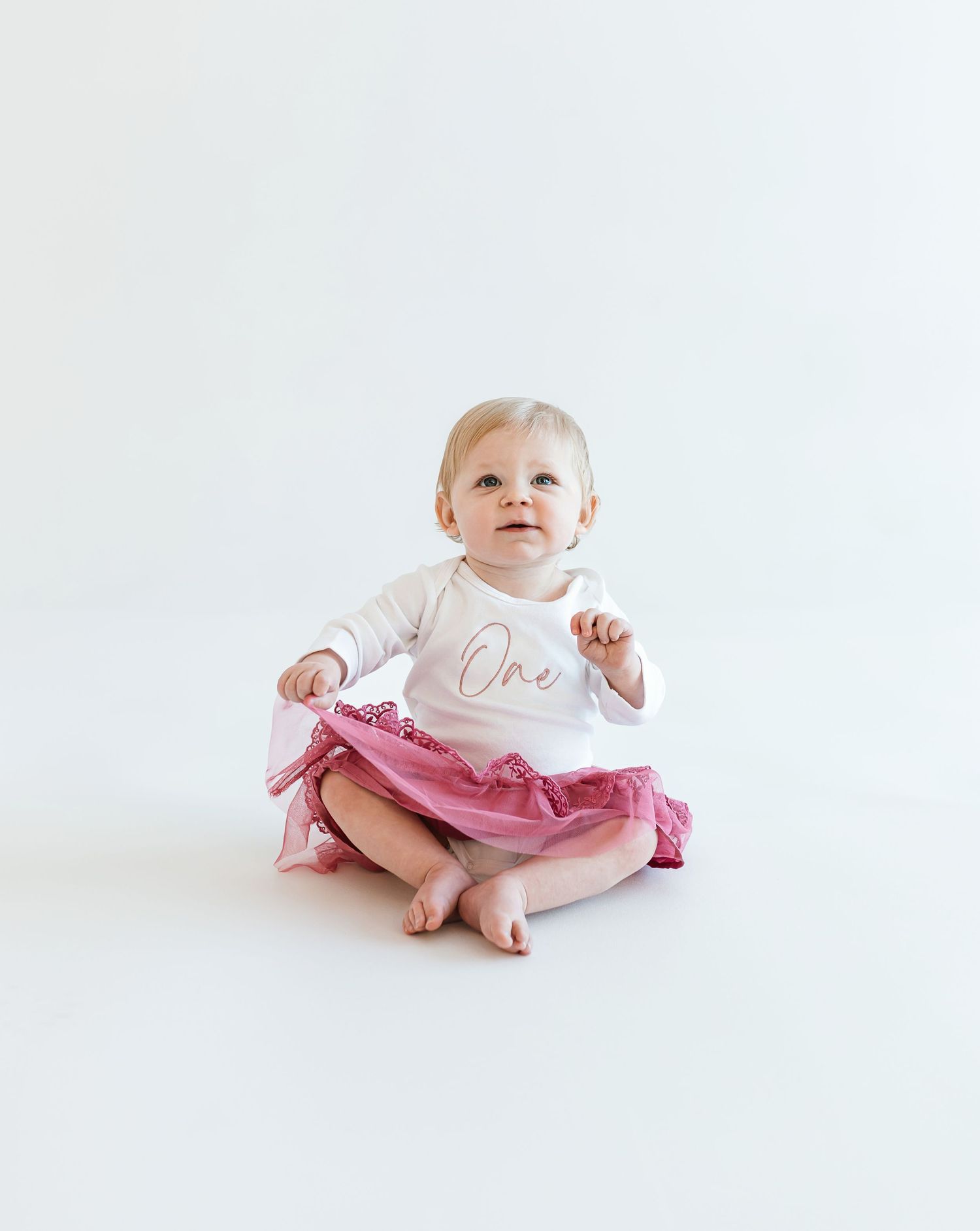 Baby wearing white shirt and pink tulle skirt sits on white background. Colorado Springs Photographer.