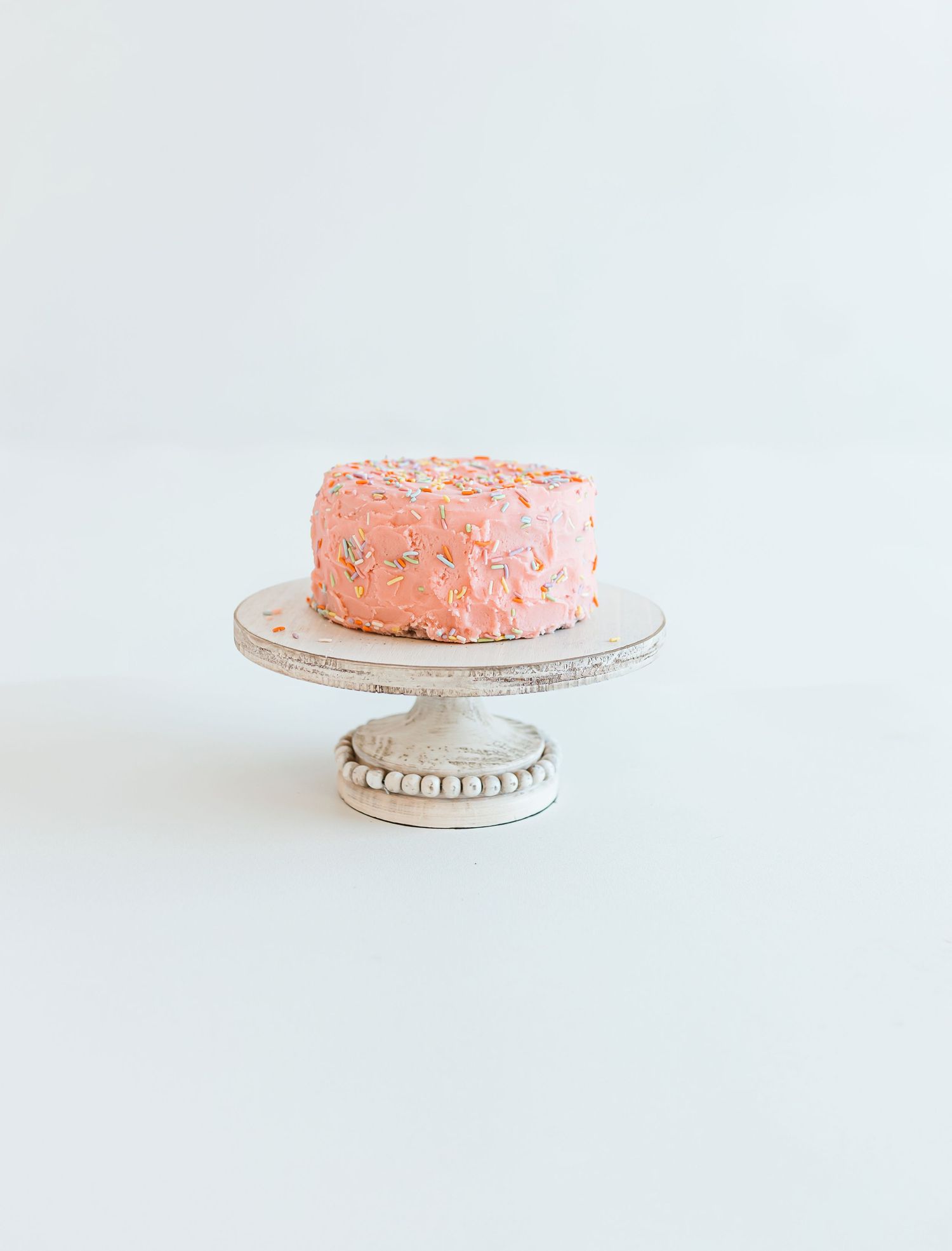 A pink frosted cake with sprinkles sits on an elegant white ceramic cake stand against a minimal white background. Colorado Springs