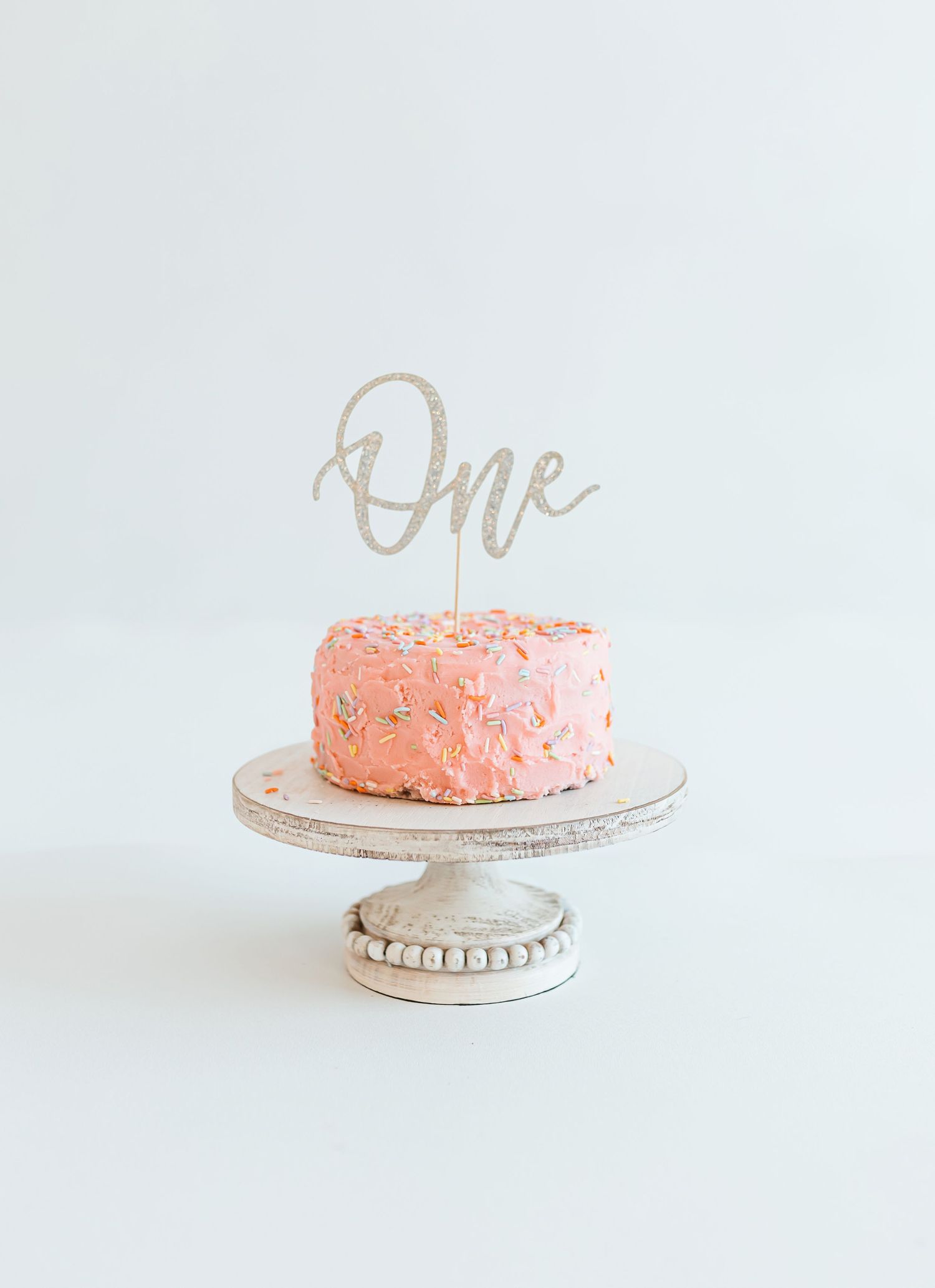 A pink birthday cake with "One" cake topper displayed on a white pedestal cake stand against white background. Colorado Springs Photographer
