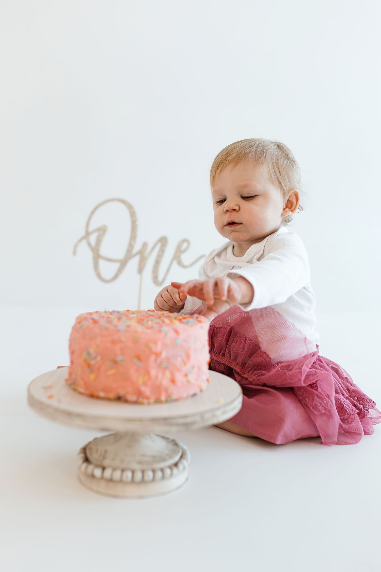 Baby in white shirt and pink pants reaches for pink birthday cake with 'One' topper on white cake stand. Colorado Springs Photographer.