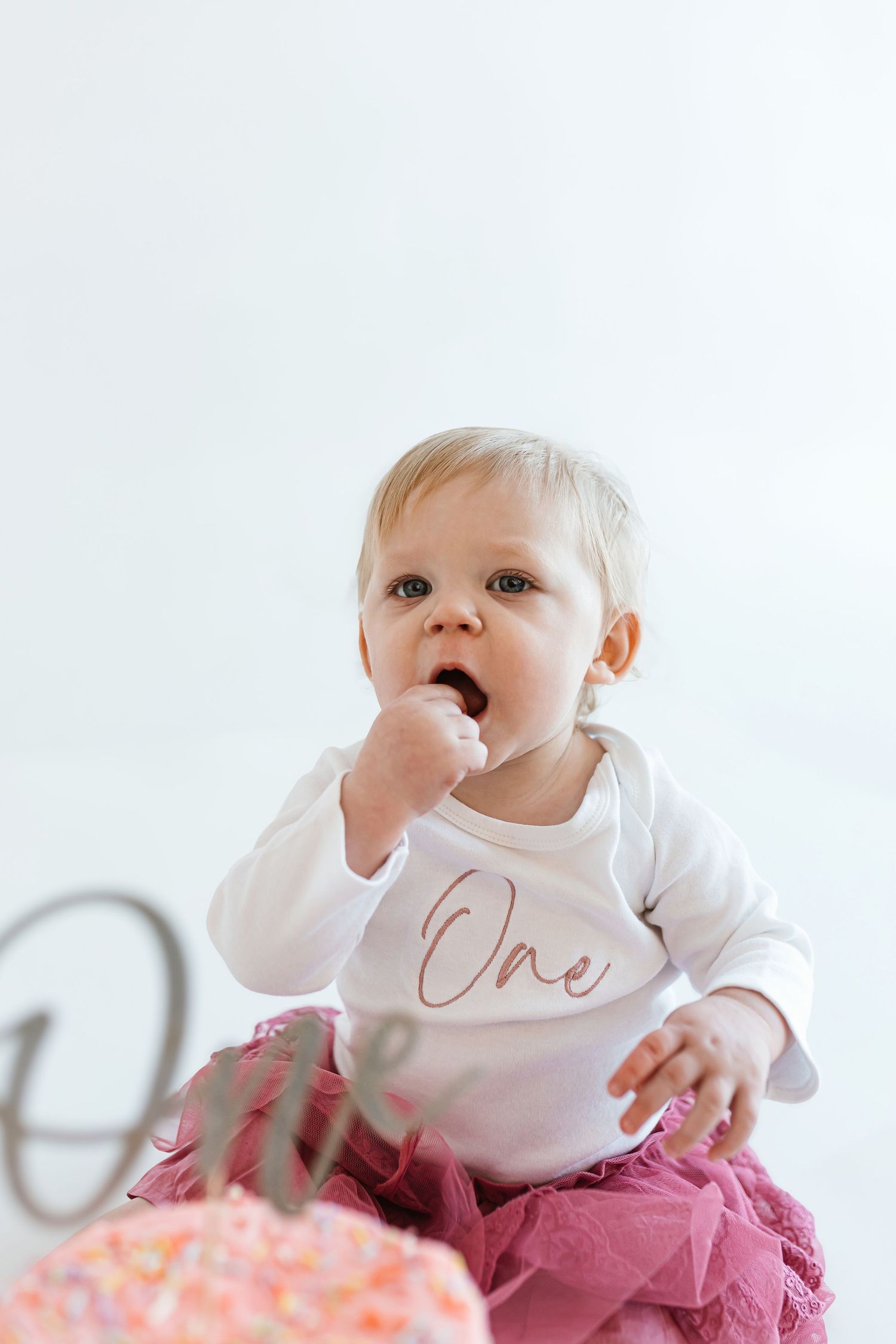 A small child in a white 'One' bodysuit and pink tutu reaching toward a birthday cake. Colorado Springs Photographer.