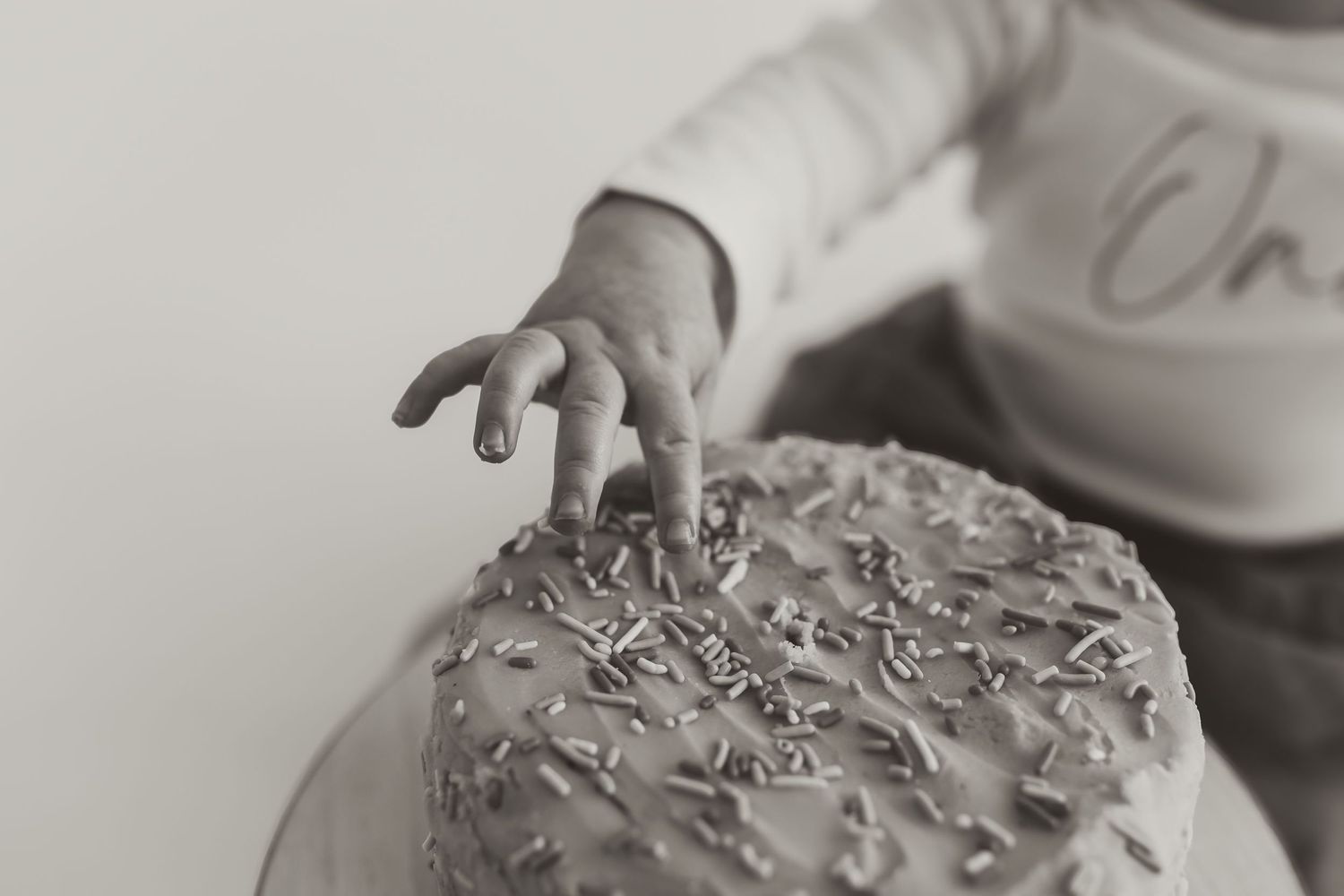 A small hand reaches out to touch the top of a frosted cake decorated with sprinkles in a black and white photograph. Colorado Springs Photo