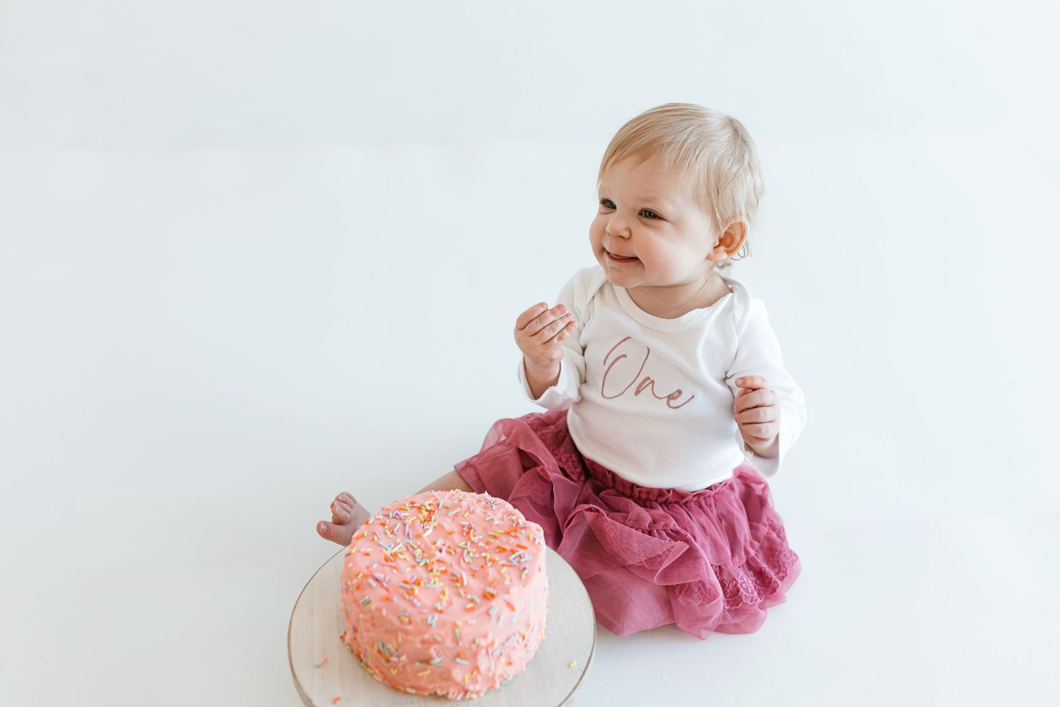 Baby in white shirt and pink skirt sits next to pink cake on white background. Colorado Springs Photographer.