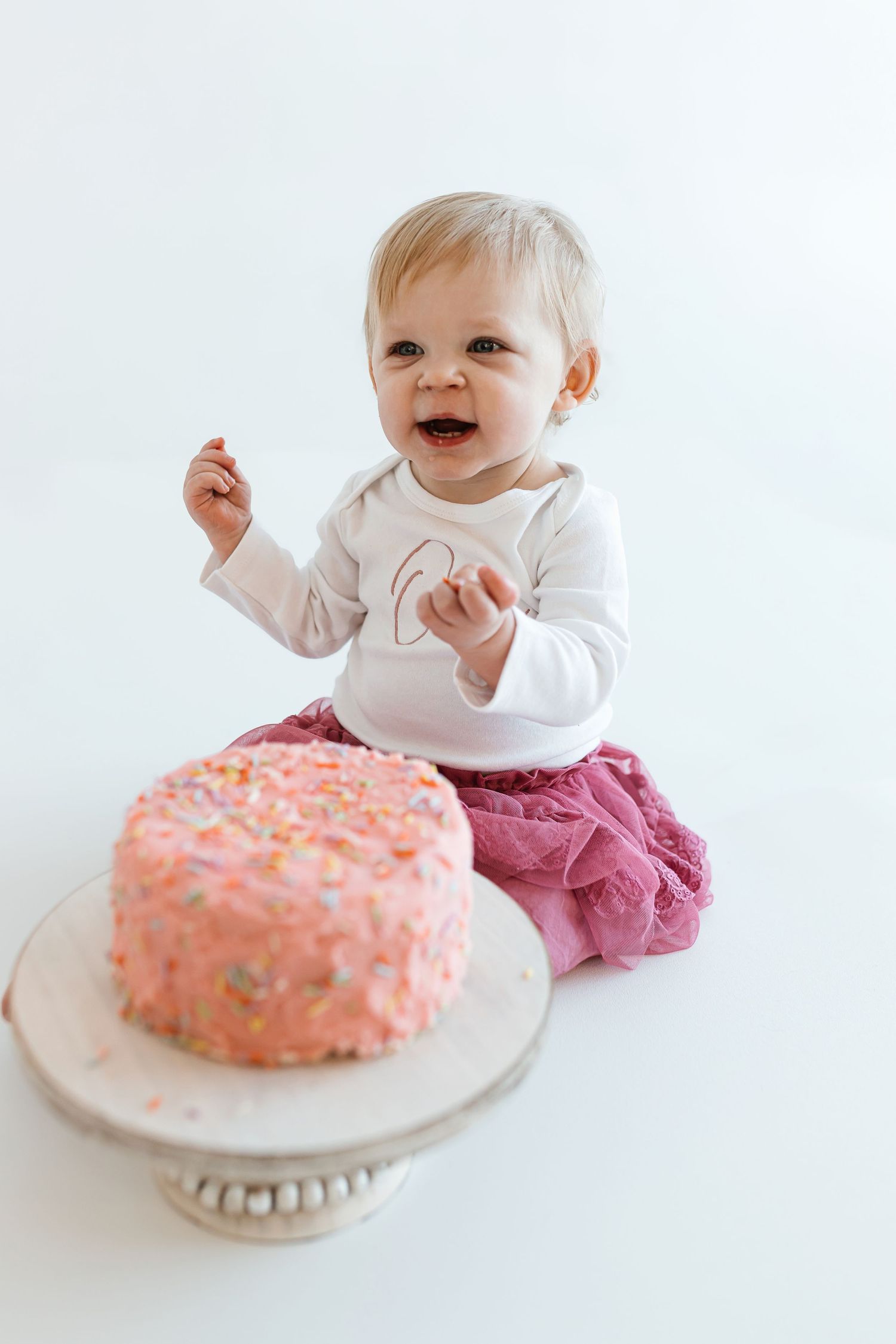 A happy baby in pink pants sits next to a pink frosted birthday cake with sprinkles on a white pedestal stand. Colorado Springs Photographer