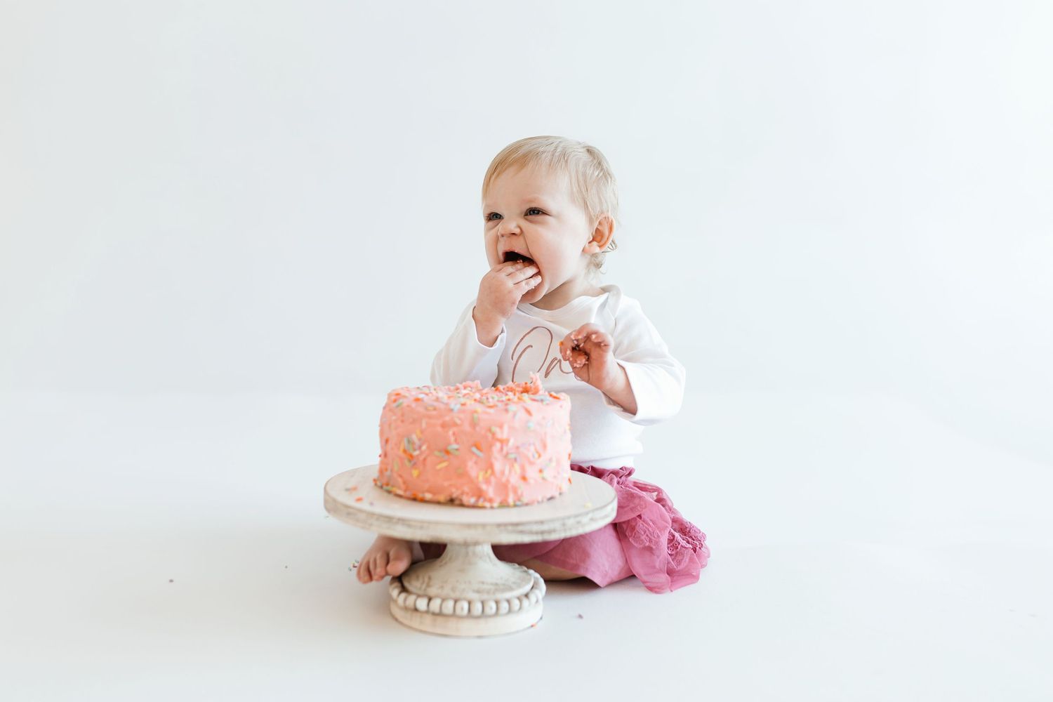 A laughing baby sits on the floor next to a pink frosted cake on a white cake stand against a white background. Colorado Springs Photograph
