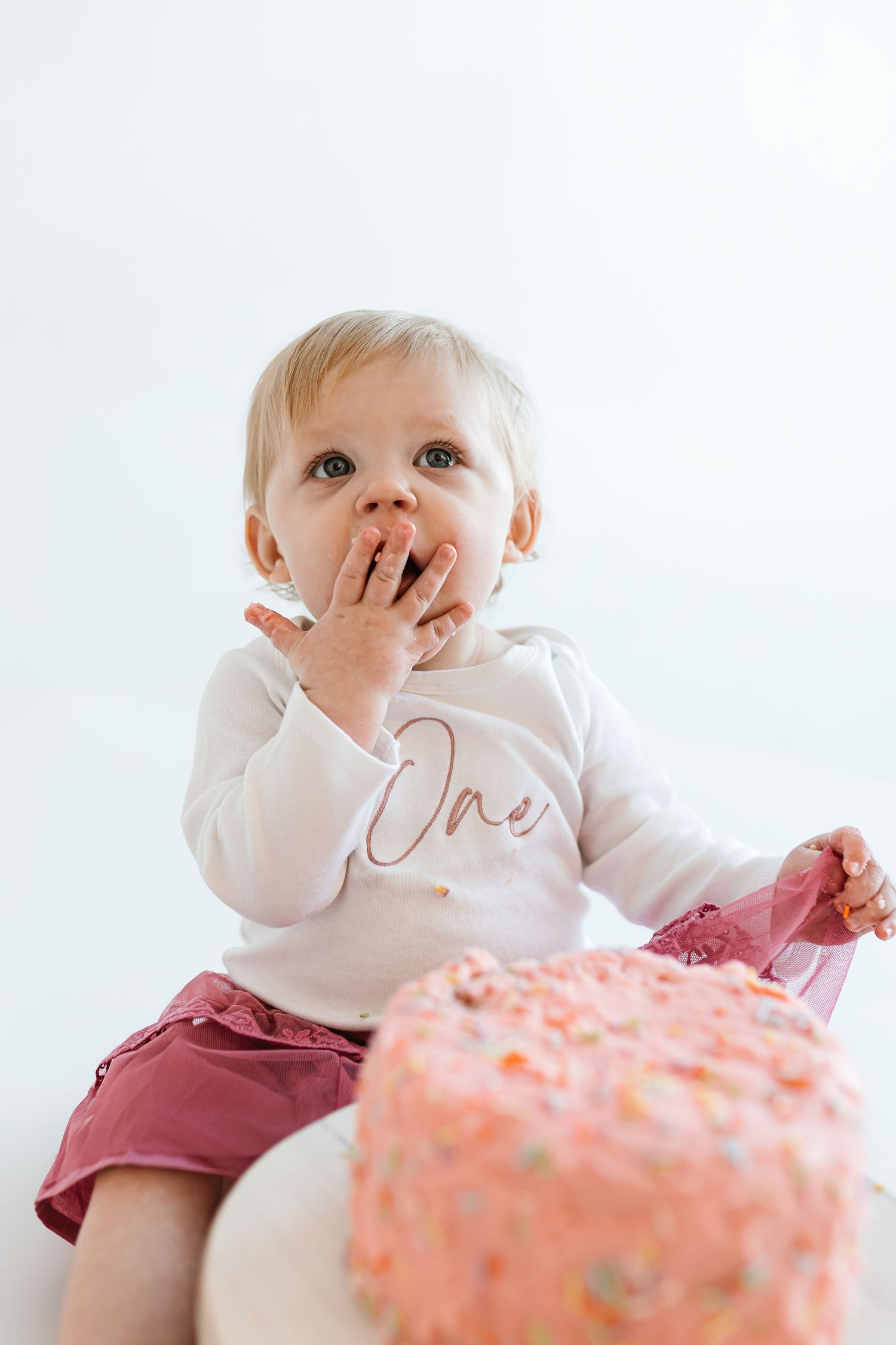 A toddler tasting cake frosting while wearing a white 'One' outfit and pink tutu. Colorado Springs Photographer.