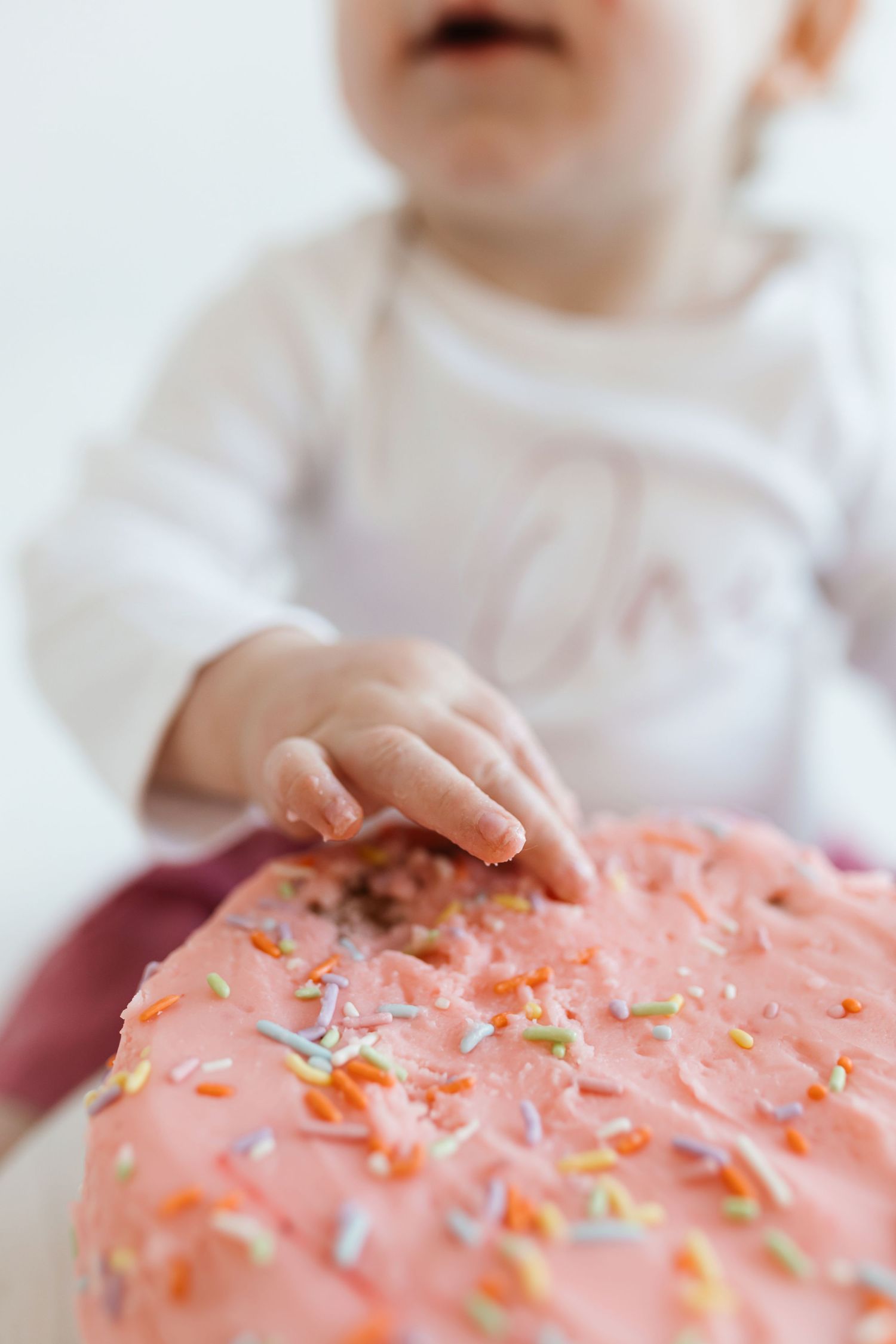 Small hands reaching to touch a pink frosted birthday cake decorated with colorful sprinkles. Colorado Springs Photographer.