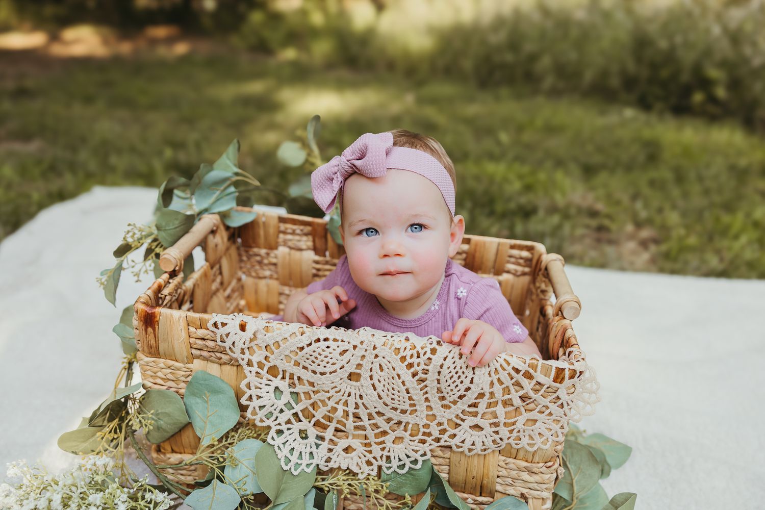 Baby in purple dress sitting in decorated wicker basket with greenery and lace details. Colorado Springs Photographer.
