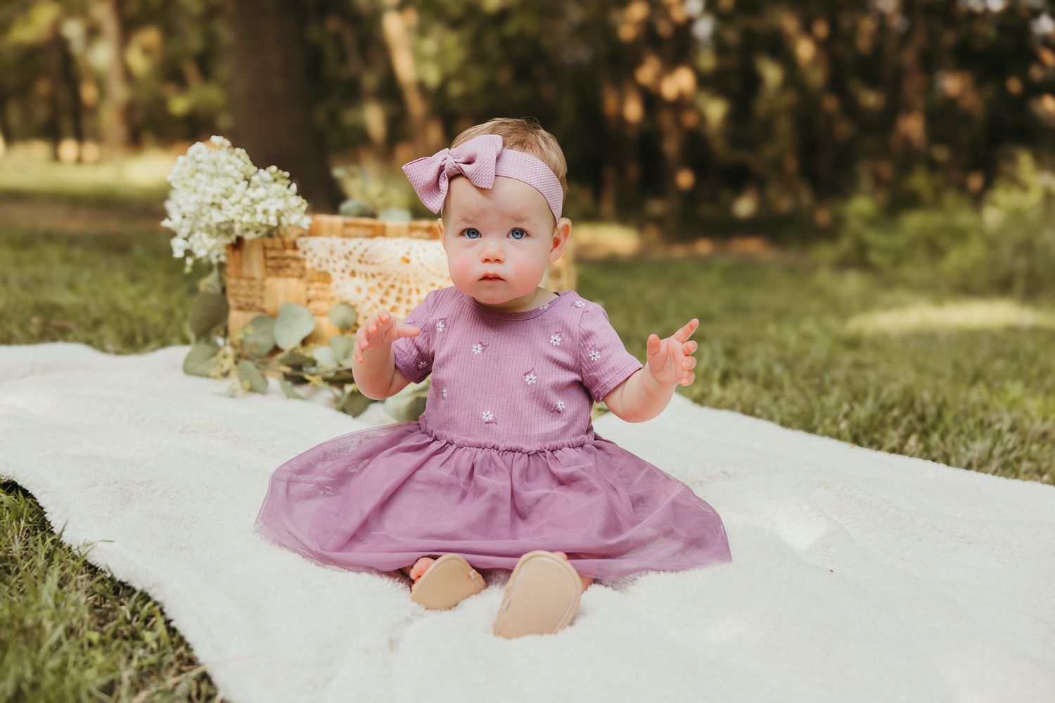 Baby in mauve dress sitting on white blanket with wicker basket and baby's breath flowers. Colorado Springs Photographer.