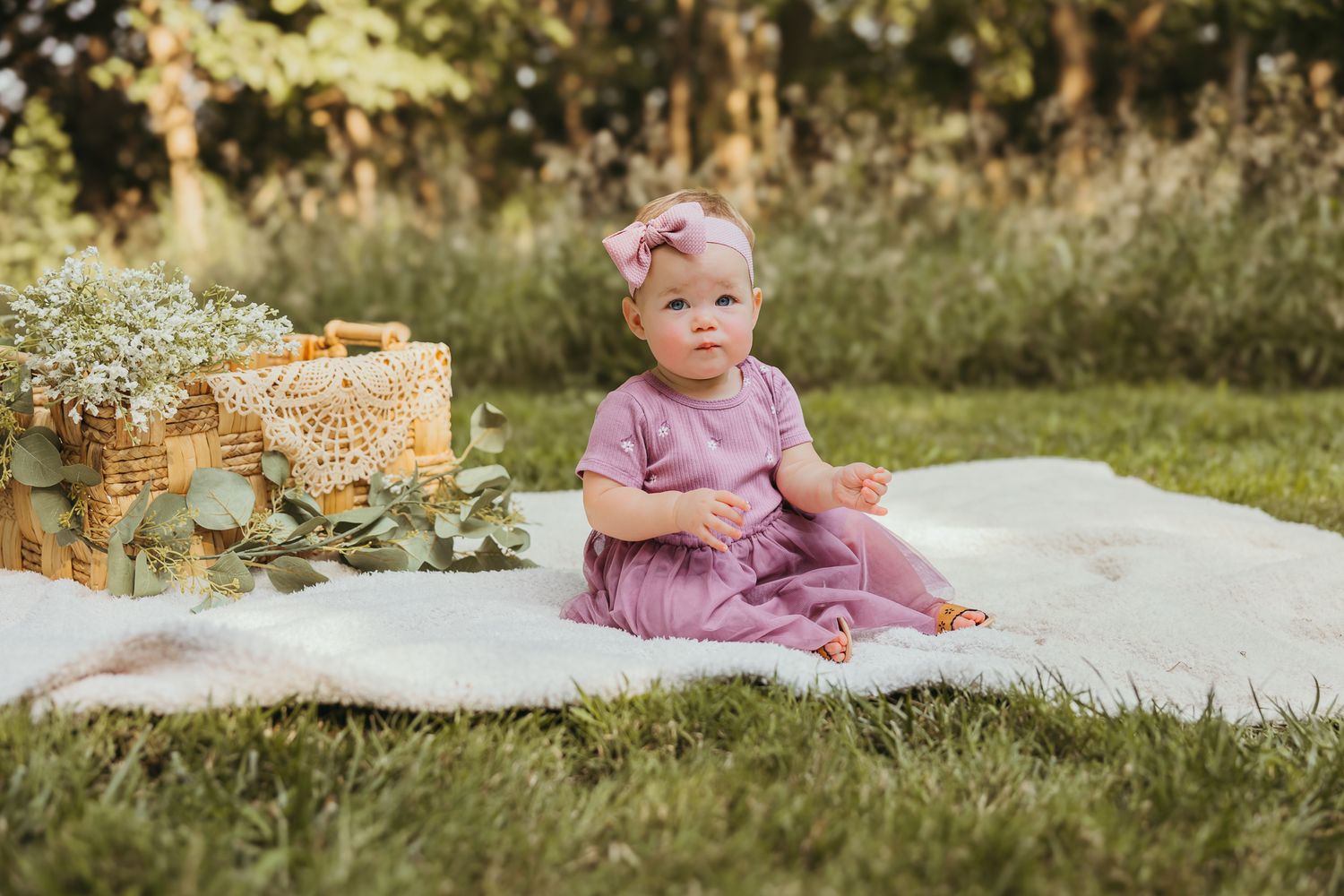 Baby in dusty rose dress sitting on white blanket with wicker basket and flowers. Colorado Springs Photographer.