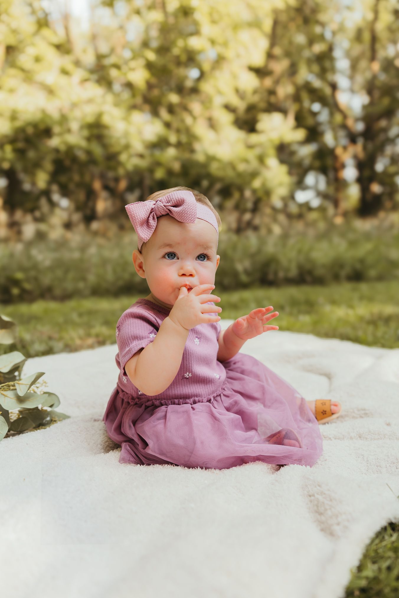 Baby in purple dress sitting on white blanket outdoors with finger in mouth. Colorado Springs Photographer.