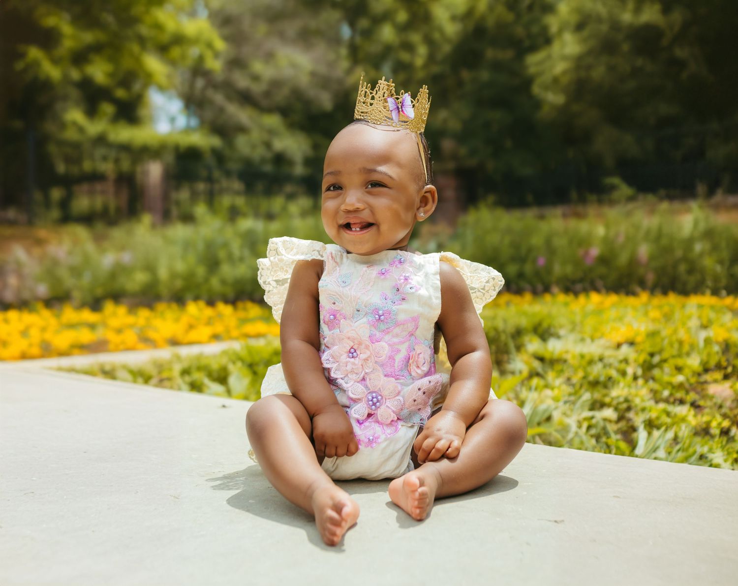 Joyful baby in floral romper and crown sitting in a sunny garden with yellow flowers. Colorado Springs Photographer.