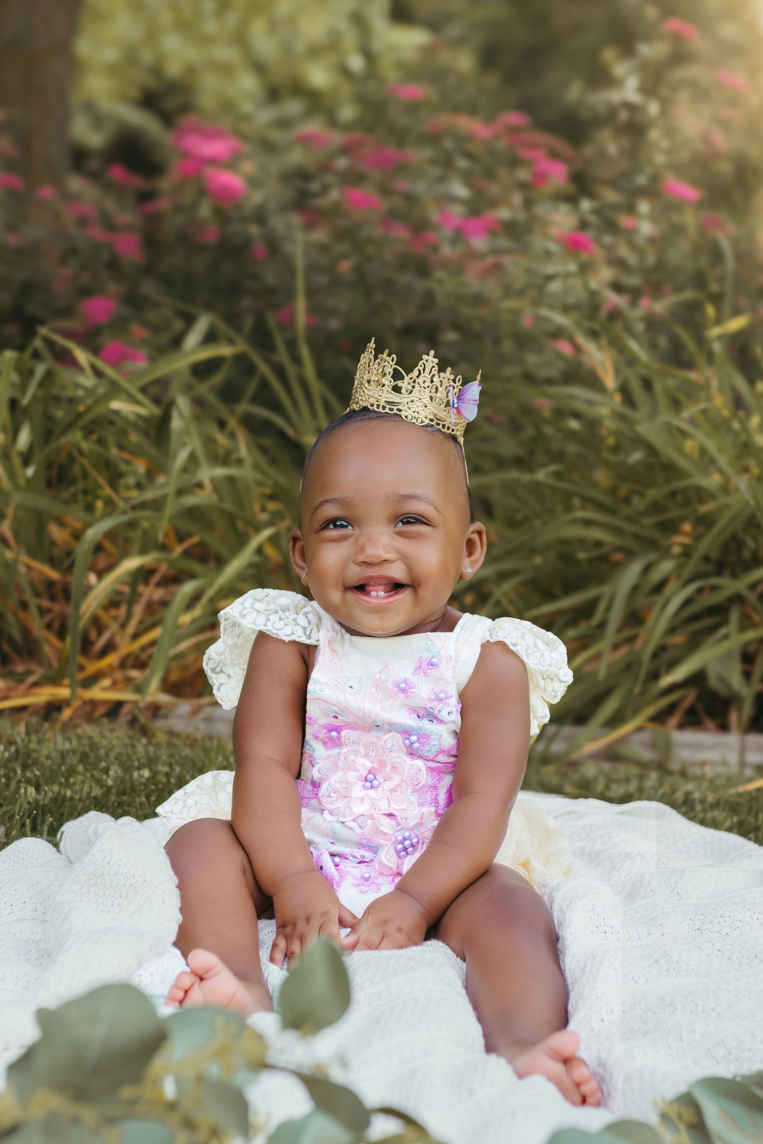 A joyful baby wearing a gold crown and floral romper sits on a white blanket in a garden with pink flowers. Colorado Springs Photographer.