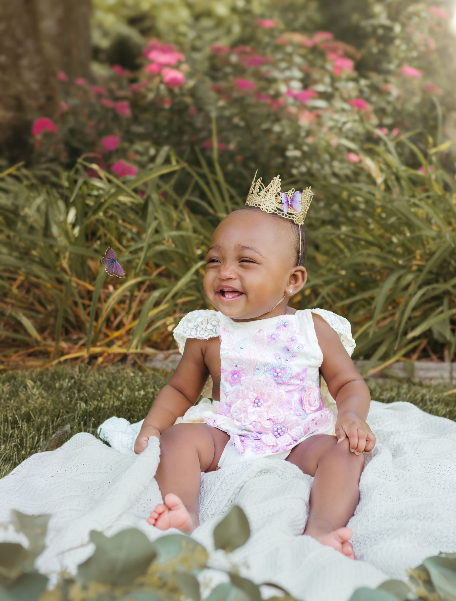 A happy baby in a lace-trimmed outfit and crown laughs while sitting outdoors with pink flowers in background. Colorado Springs Photographer