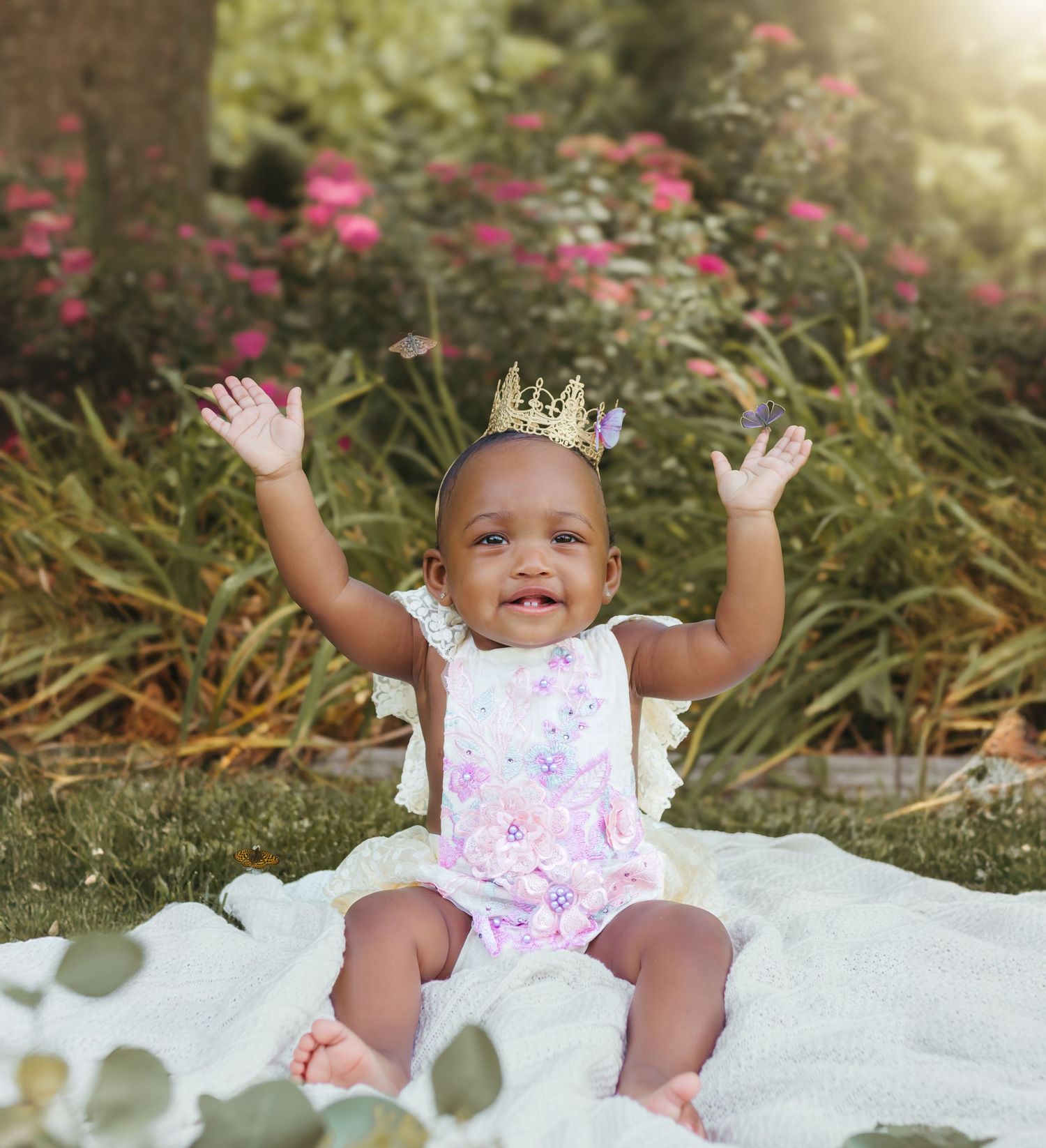 A cheerful baby in a pink romper and crown raises their arms in celebration while sitting on white fabric. Colorado Springs Photographer.