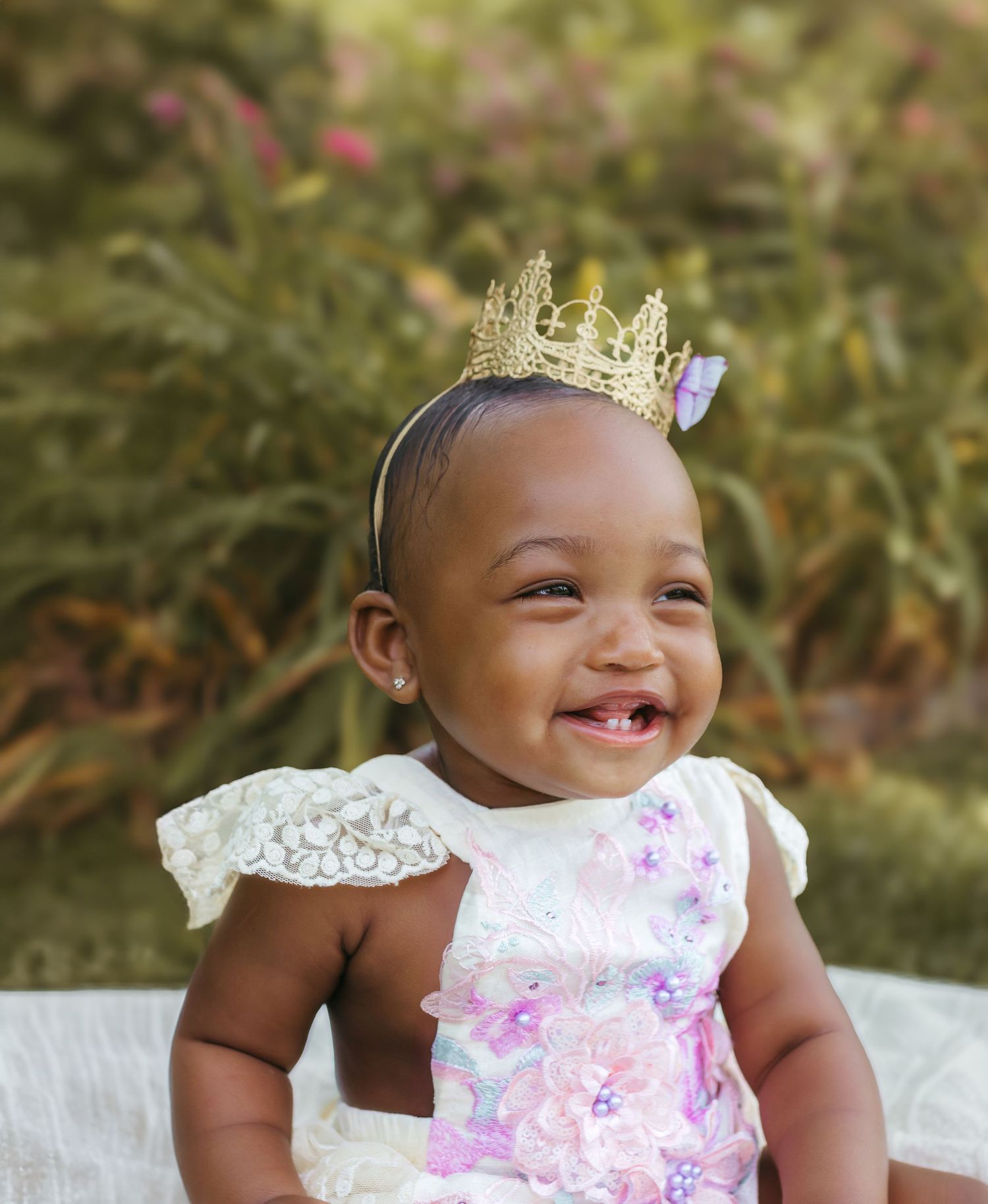A delighted baby in a floral outfit and golden crown smiles brightly against a garden background. Colorado Springs Photographer.