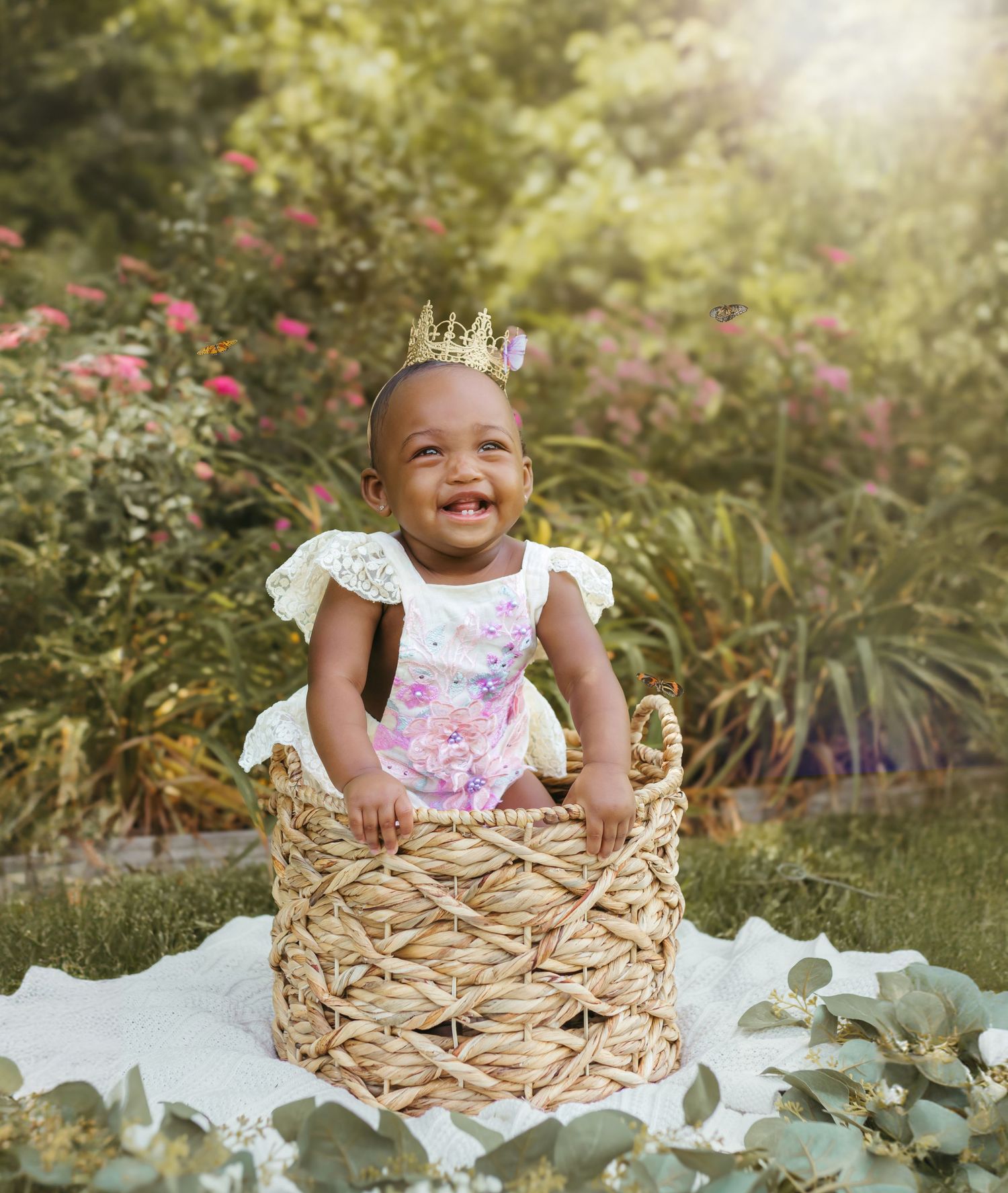 A gleeful baby wearing a crown and lace outfit sits in a woven basket on white fabric outdoors. Colorado Springs Photographer.