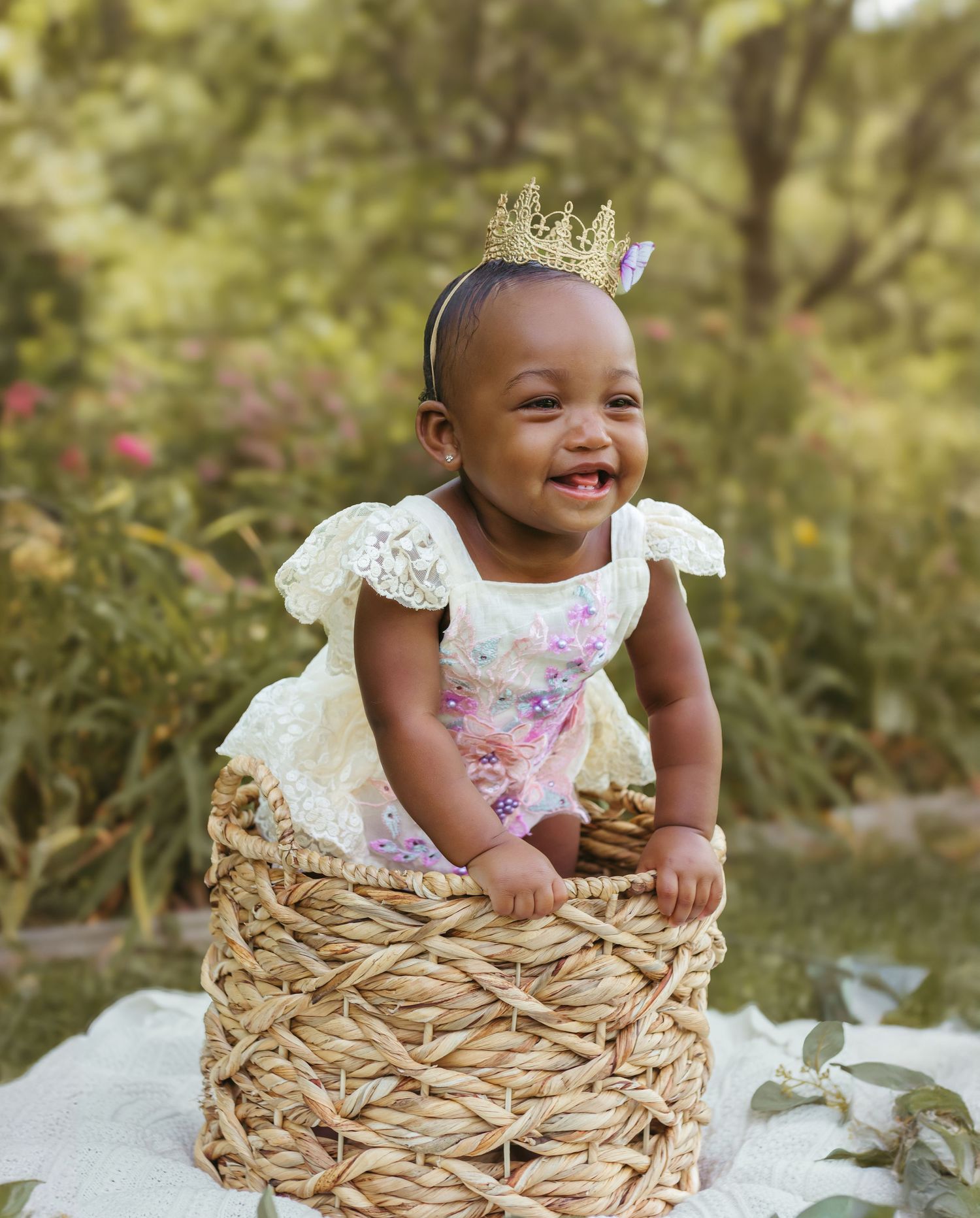 A smiling baby in a crown and floral dress poses in a woven basket with garden flowers behind. Colorado Springs Photographer.