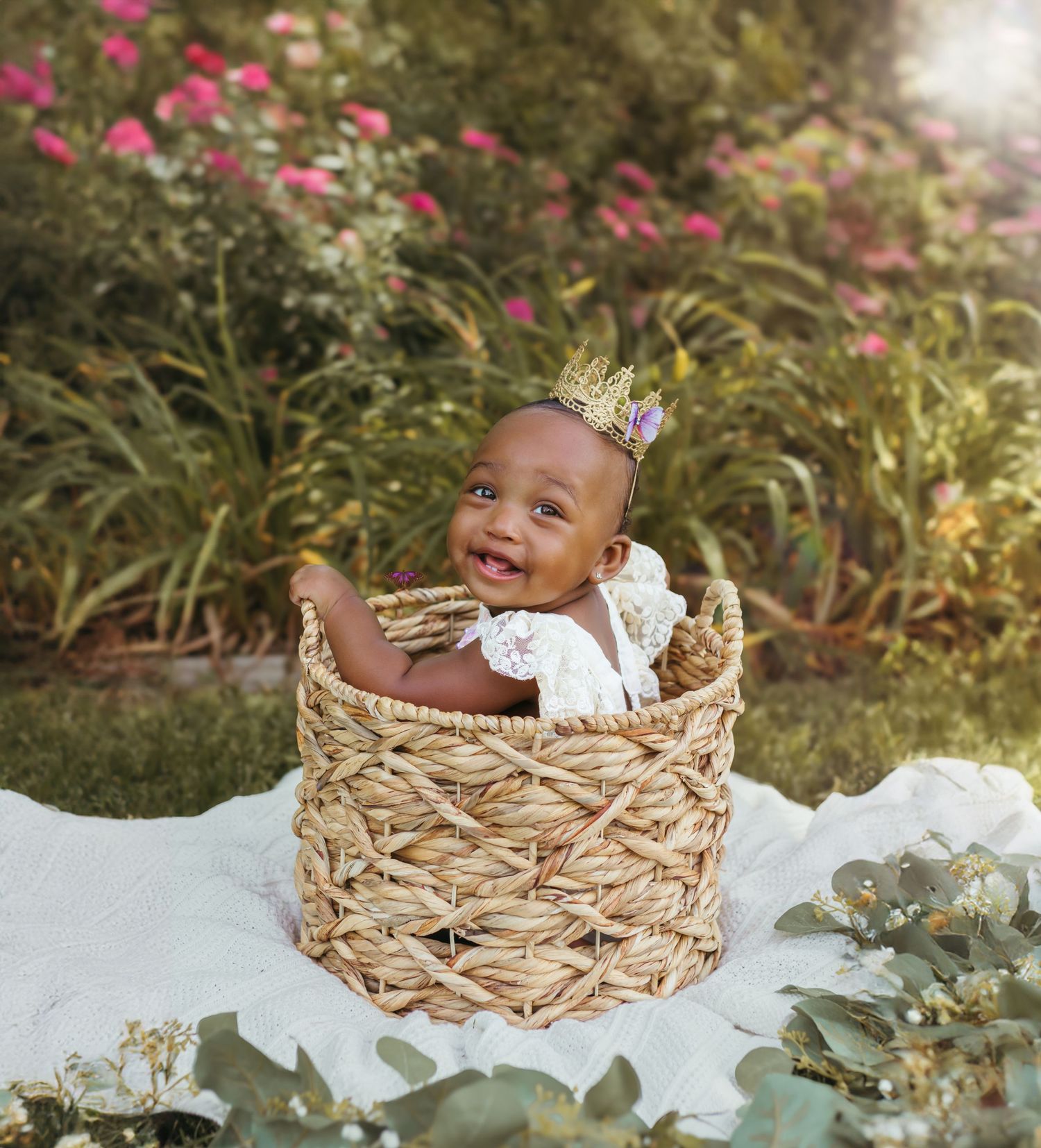 A happy baby wearing a crown and white lace outfit sits in a decorative woven basket outdoors. Colorado Springs Photographer.