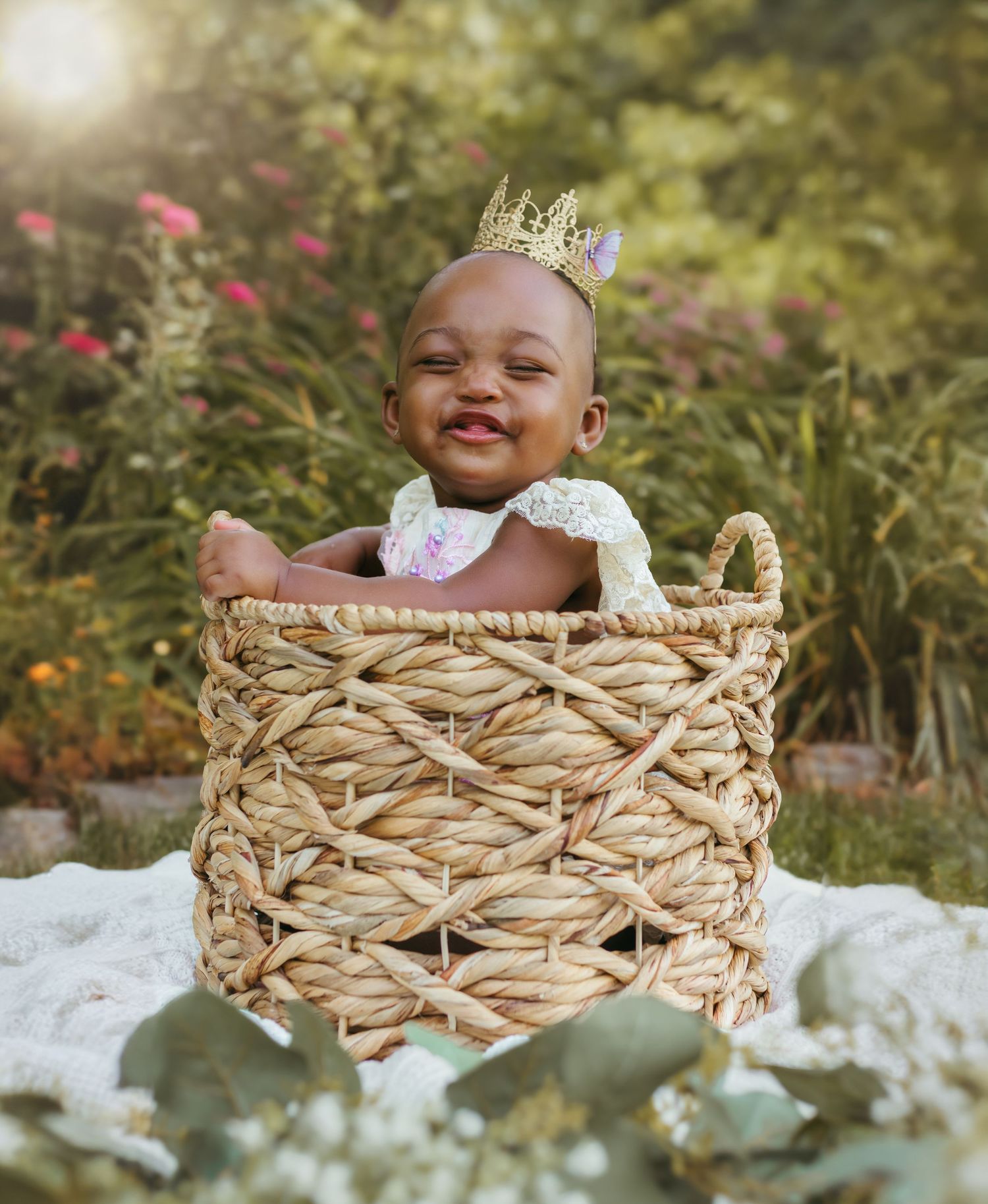 A joyful baby in a crown and dress sits in a woven basket against a backdrop of pink garden flowers. Colorado Springs Photographer.