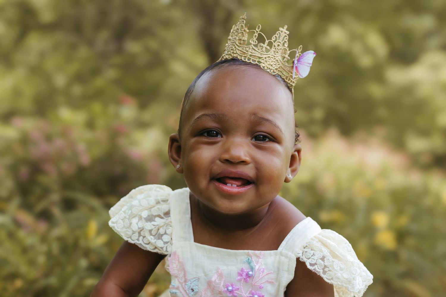 A beaming baby in a delicate dress and crown poses against blurred garden foliage. Colorado Springs Photographer.