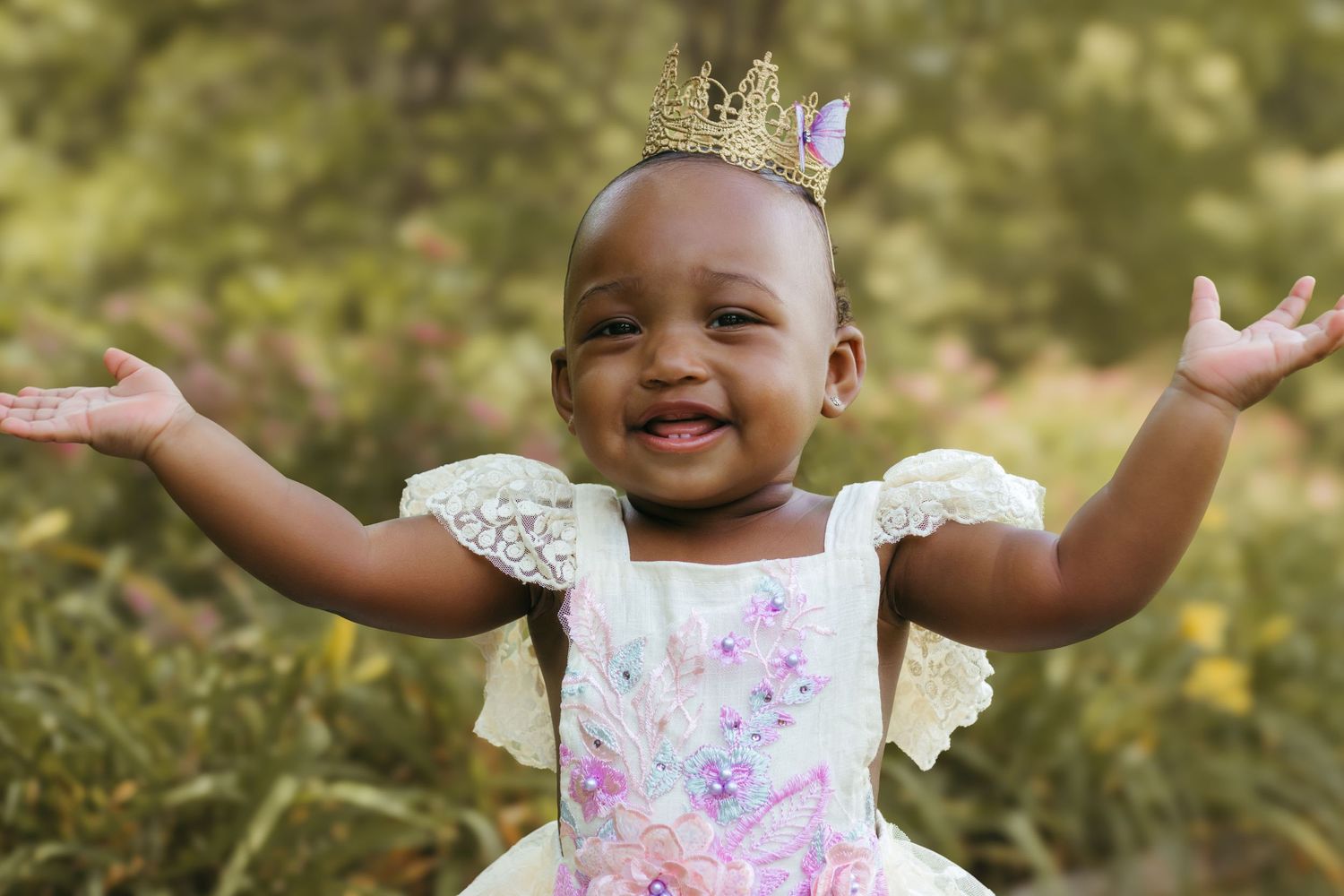 A happy baby in a crown and floral dress raises their arms joyfully against a sunny garden backdrop. Colorado Springs Photographer.