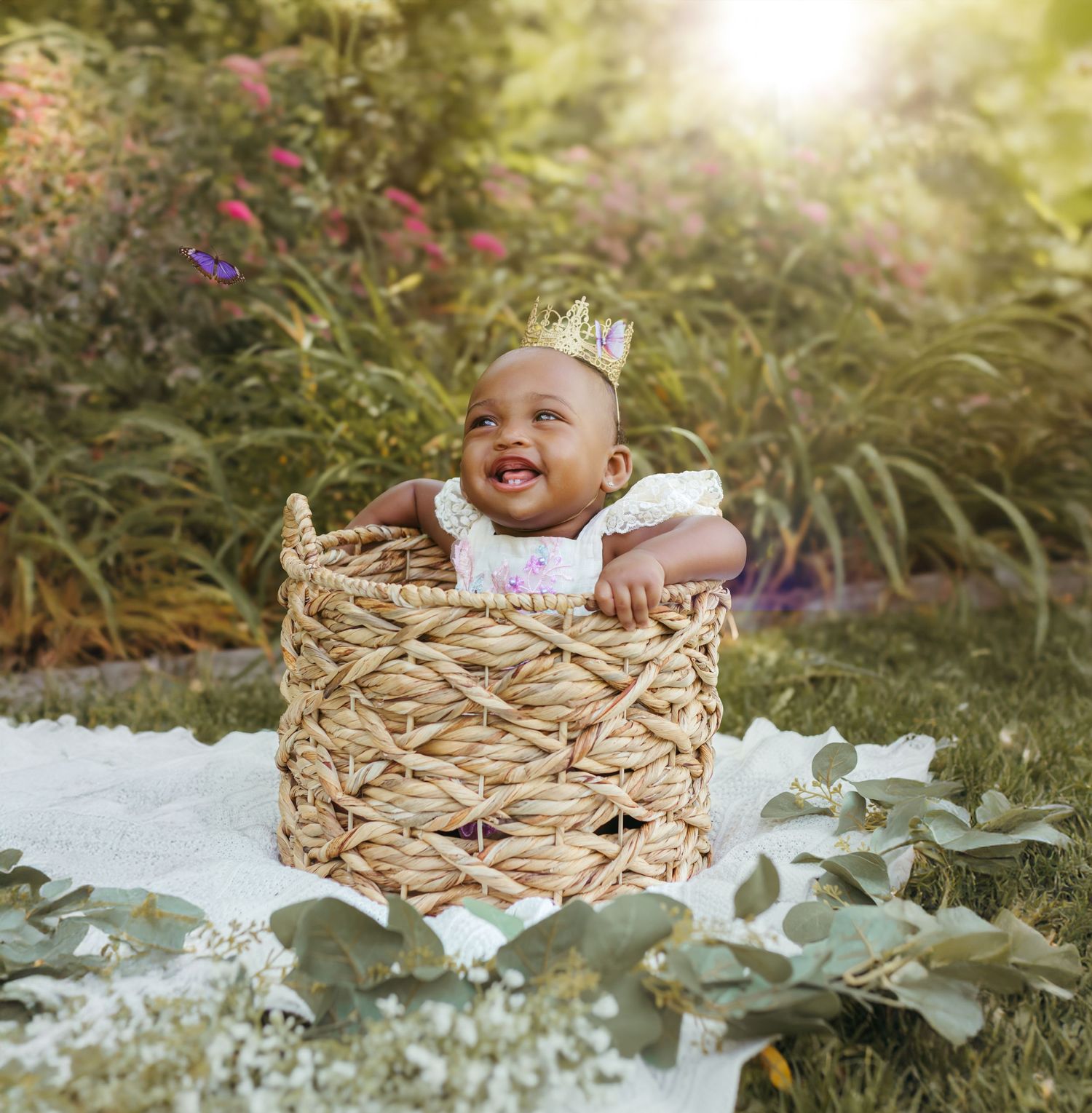 A joyful baby wearing a crown sits in a woven basket surrounded by greenery and pink flowers in a garden setting. Colorado Springs Photograp