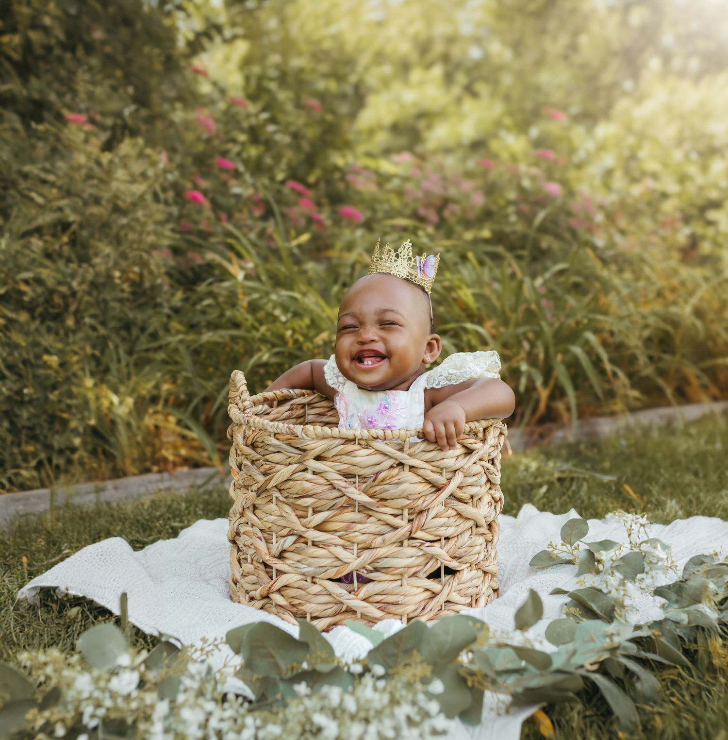 A laughing infant with a gold crown sits in a woven basket against a floral garden backdrop. Colorado Springs Photographer.