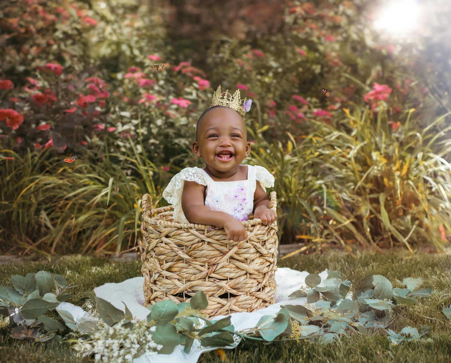 A smiling baby in white clothing and a crown poses in a rustic basket with decorative leaves around it. Colorado Springs Photographer.
