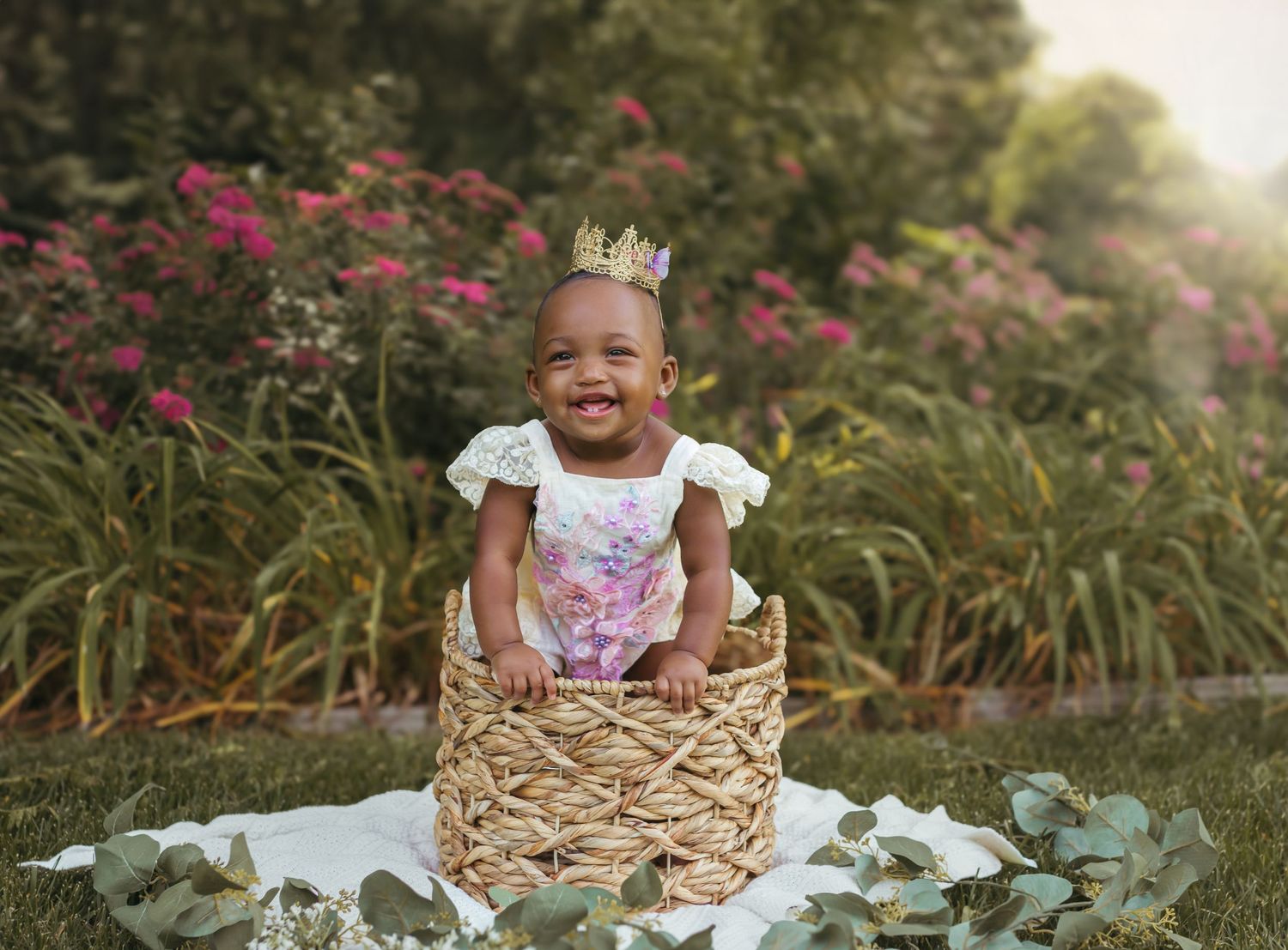 A cheerful baby in a floral dress and crown sits in a woven basket surrounded by foliage. Colorado Springs Photographer.