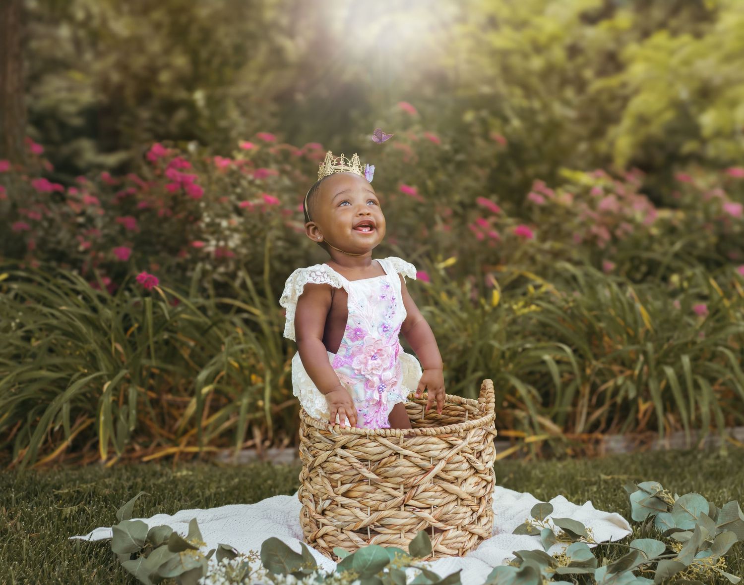 A delighted baby in white and pink attire stands in a woven basket against flowering plants. Colorado Springs Photographer.